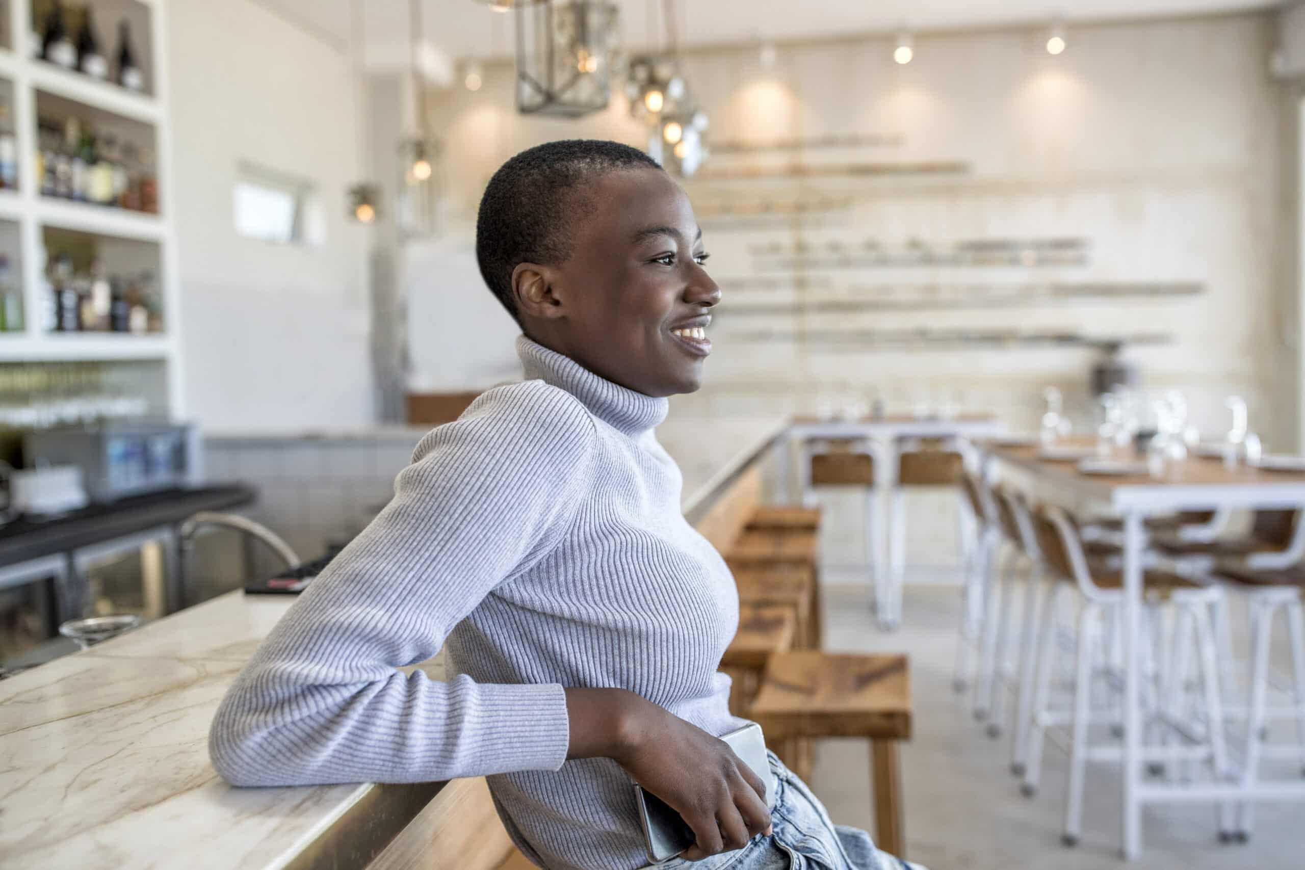 Eine fröhliche junge Frau in einem grauen Rollkragenpullover lächelt und schaut weg, während sie an einer Bar in einem modernen, hell erleuchteten Café mit stilvoller Einrichtung sitzt. © Fotografie Tomas Rodriguez