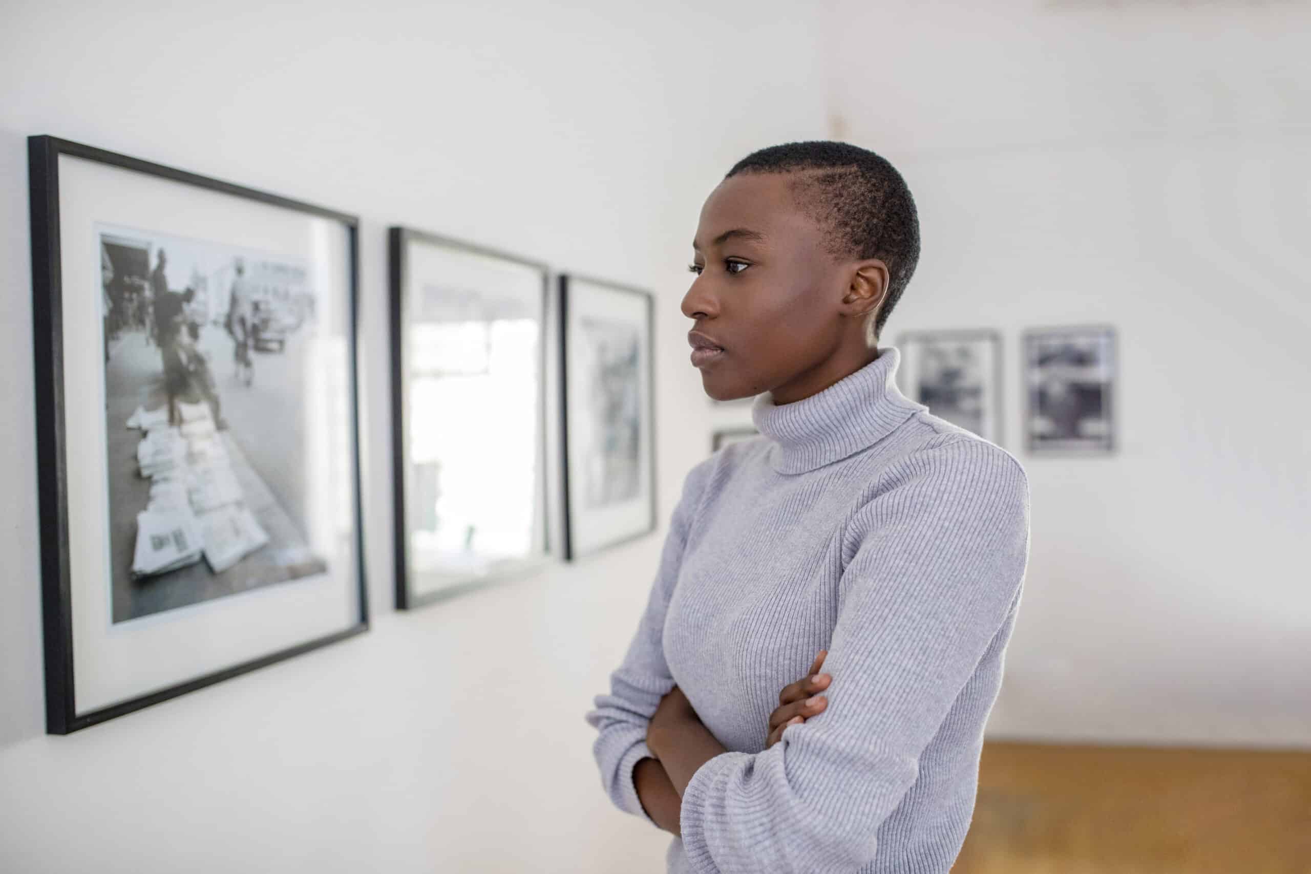 Eine junge schwarze Frau in einem hellgrauen Rollkragenpullover steht nachdenklich in einer Kunstgalerie und betrachtet gerahmte Schwarzweißfotografien an weißen Wänden. © Fotografie Tomas Rodriguez