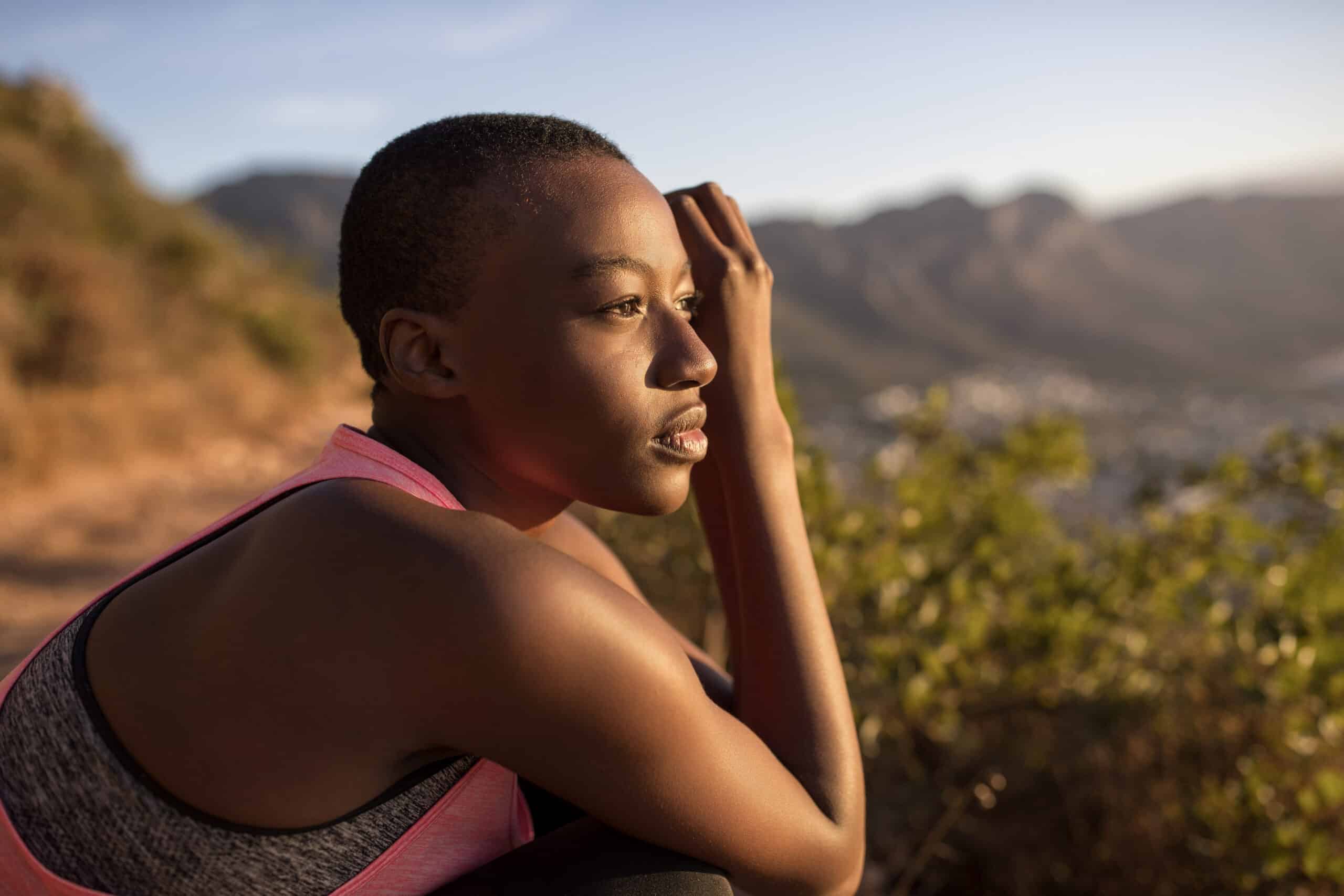 Eine junge schwarze Frau in Sportkleidung ruht sich aus und blickt während der goldenen Stunde nachdenklich auf eine malerische Berglandschaft. © Fotografie Tomas Rodriguez