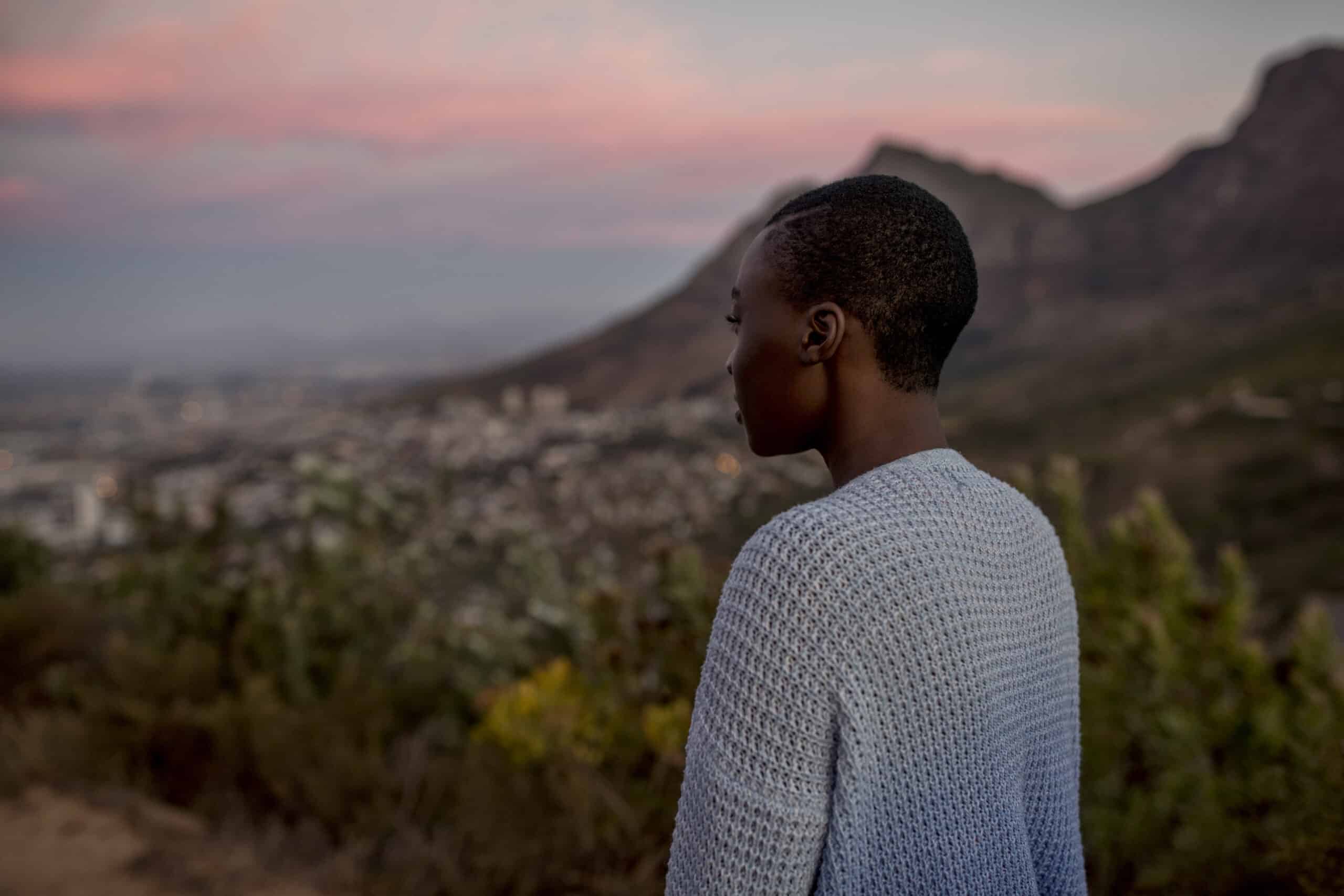 Eine Person, die in der Abenddämmerung von einem hohen Aussichtspunkt aus eine Stadtlandschaft betrachtet, mit Bergen im Hintergrund unter einem sanften rosa und blauen Himmel. © Fotografie Tomas Rodriguez