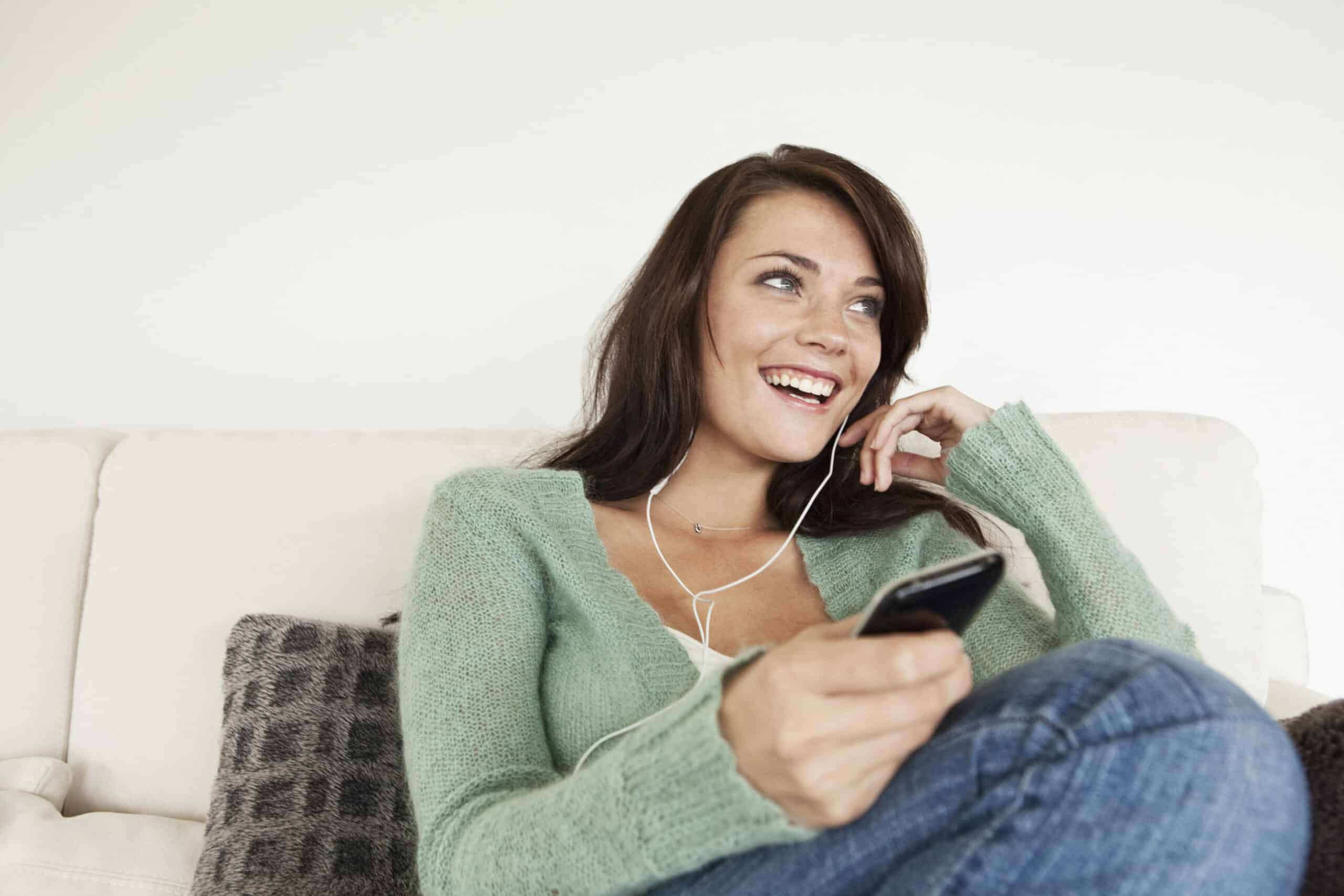 Eine Frau im grünen Pullover sitzt lächelnd auf einem Sofa, hält ein Smartphone in der Hand und trägt Kopfhörer. Sie scheint ein Gespräch zu genießen oder etwas zu hören. © Fotografie Tomas Rodriguez
