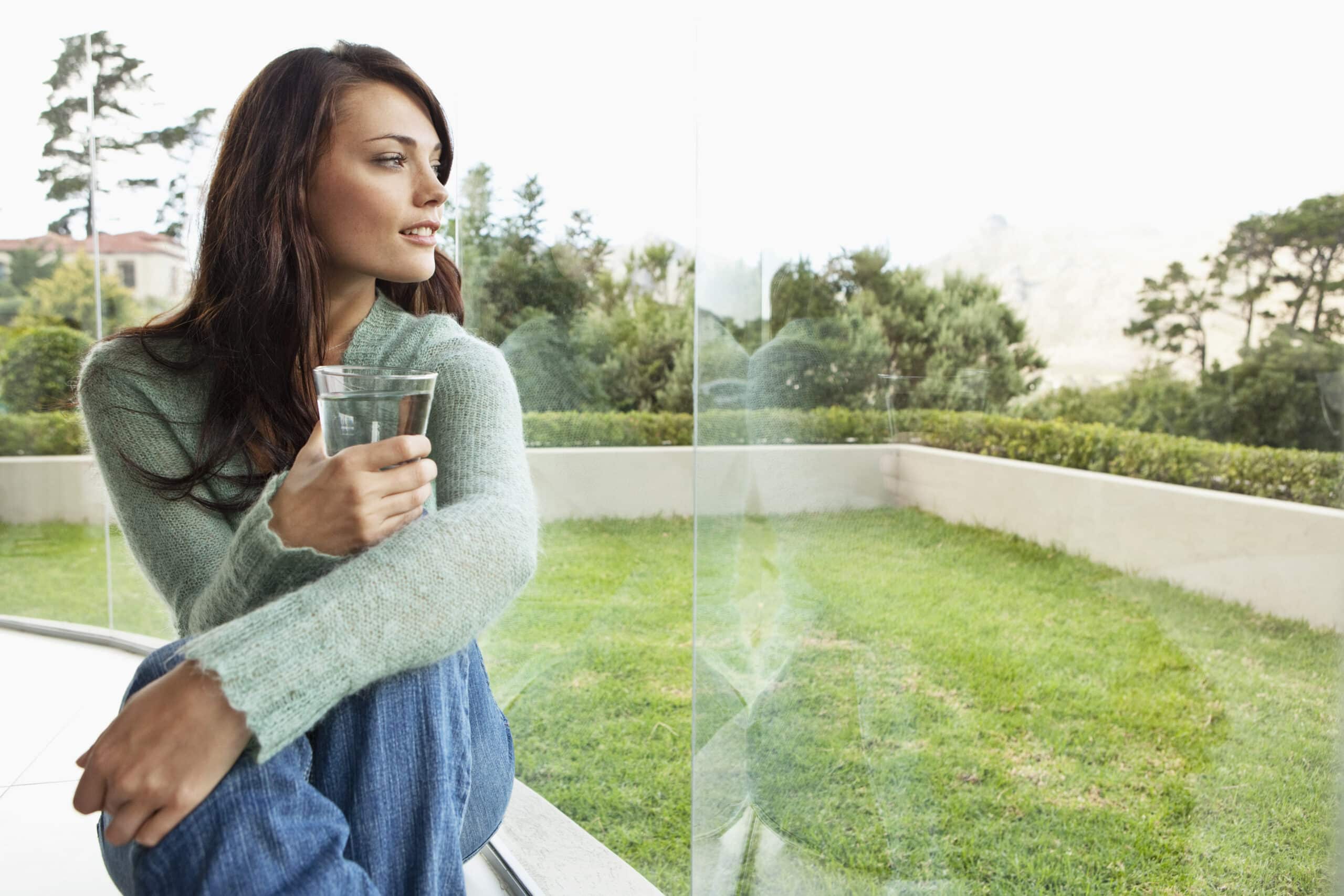 Eine Frau in einem kuscheligen Pullover hält ein Glas Wasser, während sie an einem Balkongeländer lehnt und auf einen üppigen Garten mit reflektierendem Glas neben sich blickt. © Fotografie Tomas Rodriguez