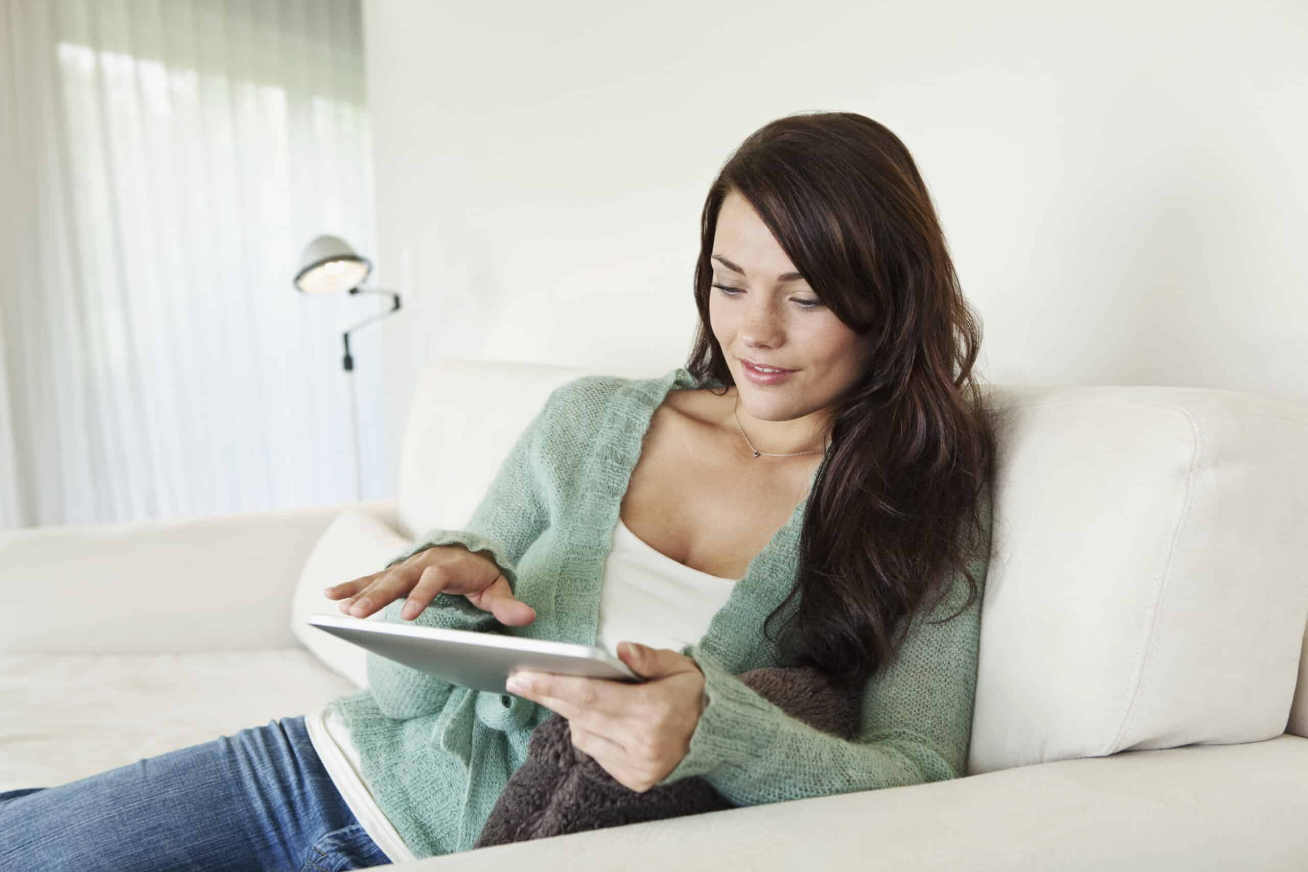 Eine Frau in einem kuscheligen grünen Pullover sitzt mit einem Tablet-PC auf einem weißen Sofa. Sie befindet sich in einem Innenbereich mit einer hellen, luftigen Kulisse aus transparenten Vorhängen. © Fotografie Tomas Rodriguez