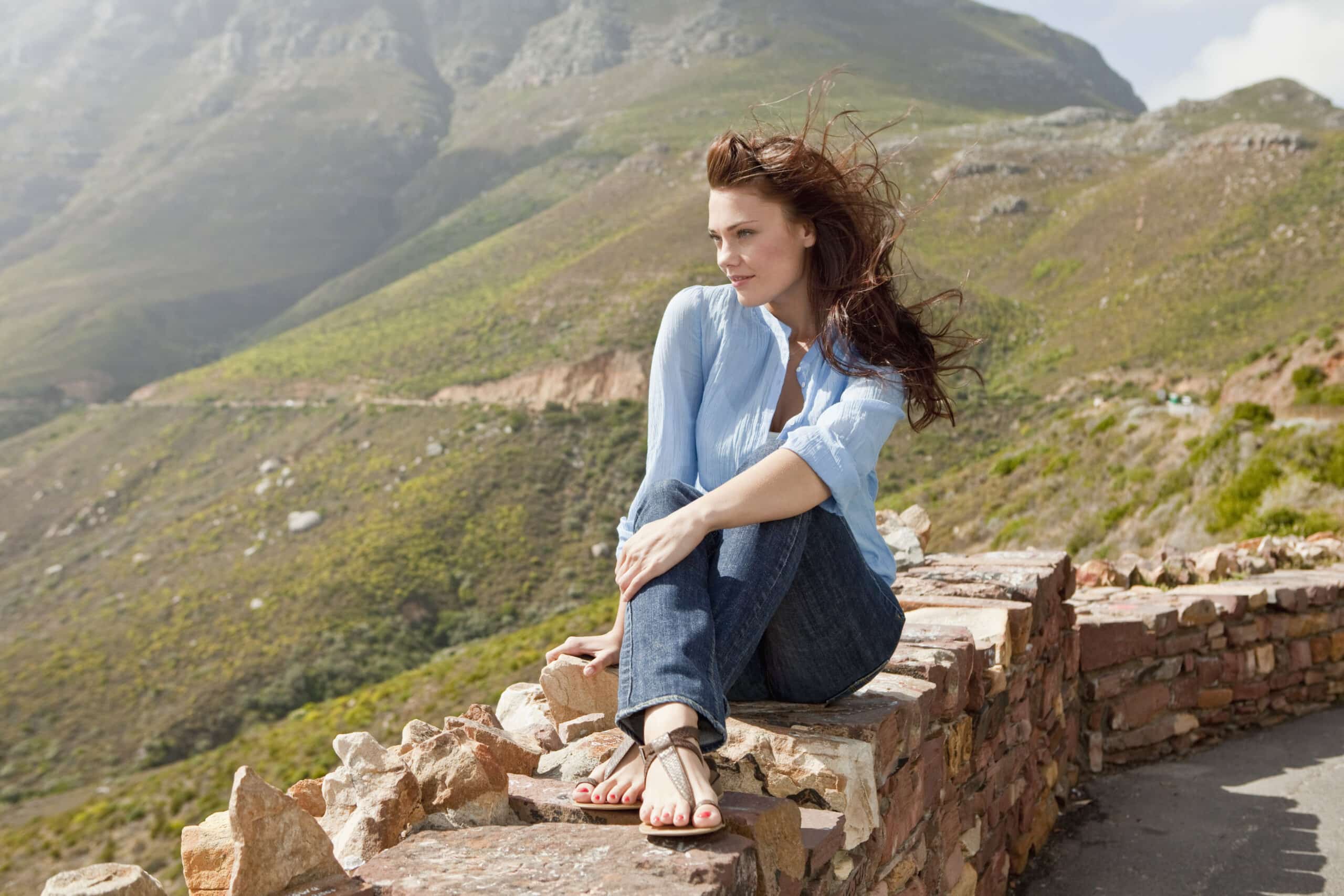 Eine Frau in blauem Hemd und Jeans sitzt auf einer Steinmauer und blickt auf eine Berglandschaft. Ihr Haar weht im Wind. © Fotografie Tomas Rodriguez