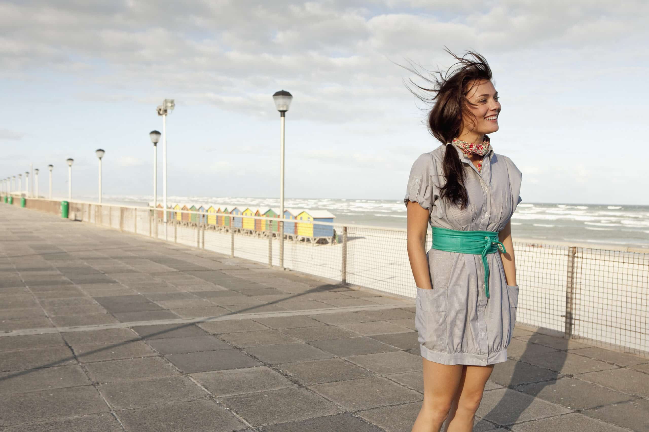 Eine fröhliche Frau steht auf einer windigen Strandpromenade, ihr Haar weht im Wind. Sie trägt ein graues Kleid mit einem grünen Gürtel und lächelt breit vor dem Hintergrund eines bewölkten Himmels und Meereswellen. © Fotografie Tomas Rodriguez