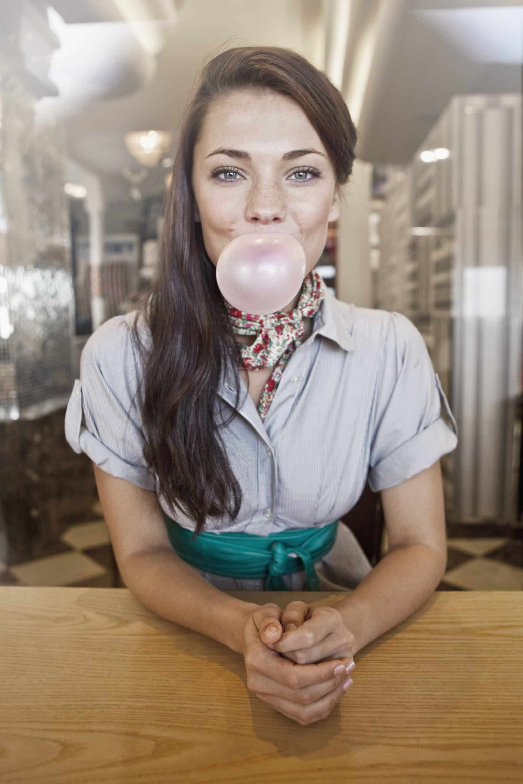 Eine Frau im Retro-Outfit sitzt in einem Diner an einem Tisch mit reflektierendem Hintergrund und lässt eine große rosa Kaugummiblase platzen. © Fotografie Tomas Rodriguez