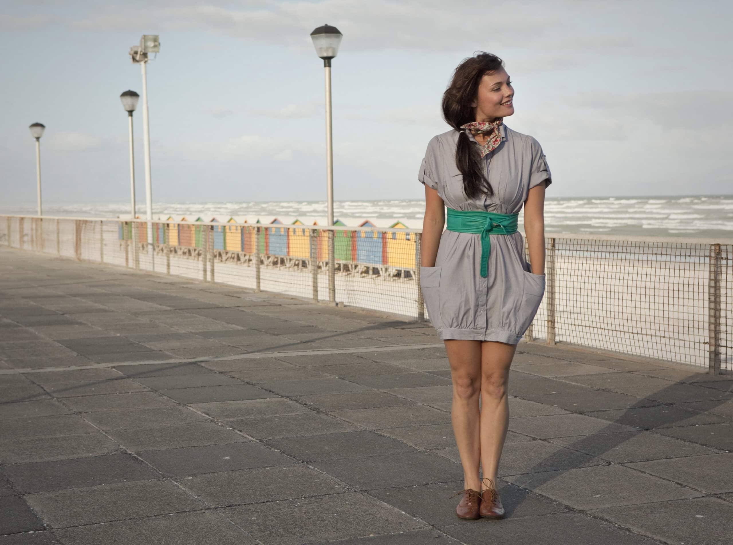Eine Frau steht auf einer Strandpromenade und blickt in die Ferne. Sie trägt ein graues Kleid mit einem grünen Gürtel und einem Schal. Im Hintergrund sind bunte Strandhütten und ein stürmisches Meer zu sehen. © Fotografie Tomas Rodriguez