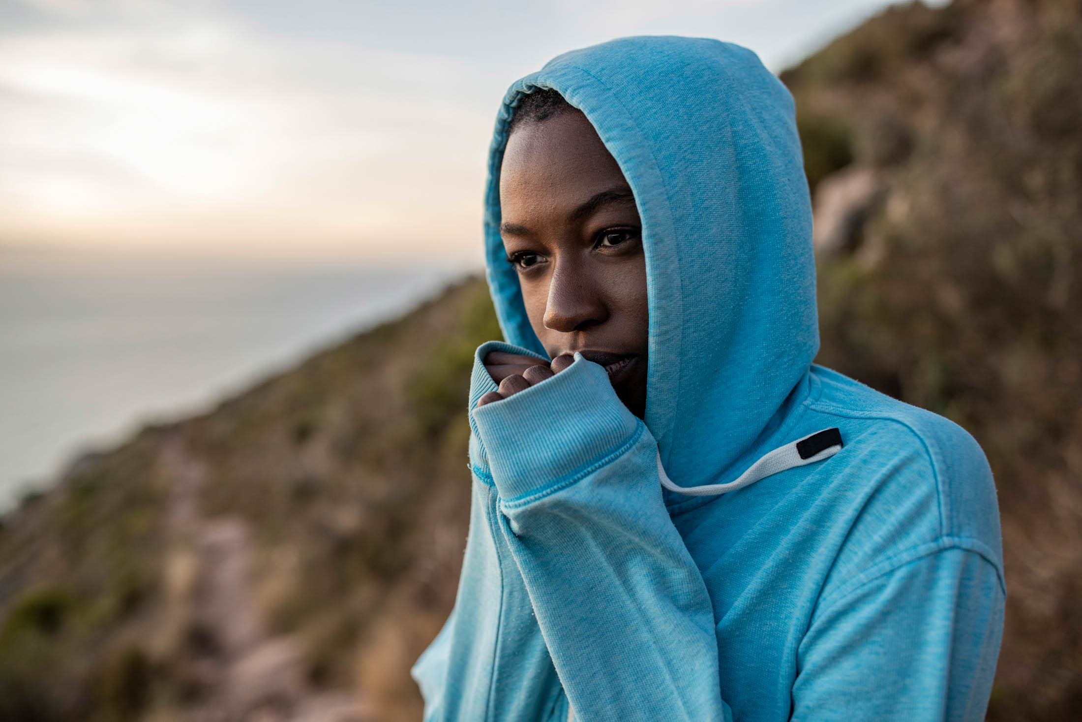 Eine Person in einem blauen Kapuzenpullover steht auf einem Berg und sieht nachdenklich aus. Im Hintergrund ist das Meer unscharf. Der Kapuzenpullover bedeckt teilweise ihren Mund. © Fotografie Tomas Rodriguez