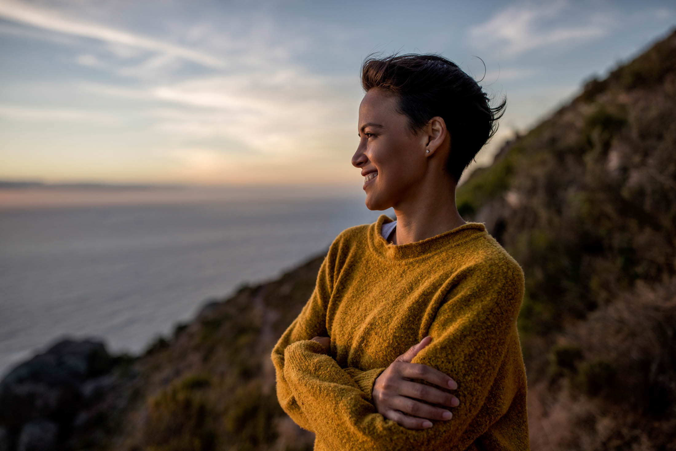 Eine fröhliche Frau in einem gelben Pullover steht bei Sonnenuntergang auf einer Küstenklippe, blickt mit heiterem Gesichtsausdruck und verschränkten Armen auf das Meer. © Fotografie Tomas Rodriguez