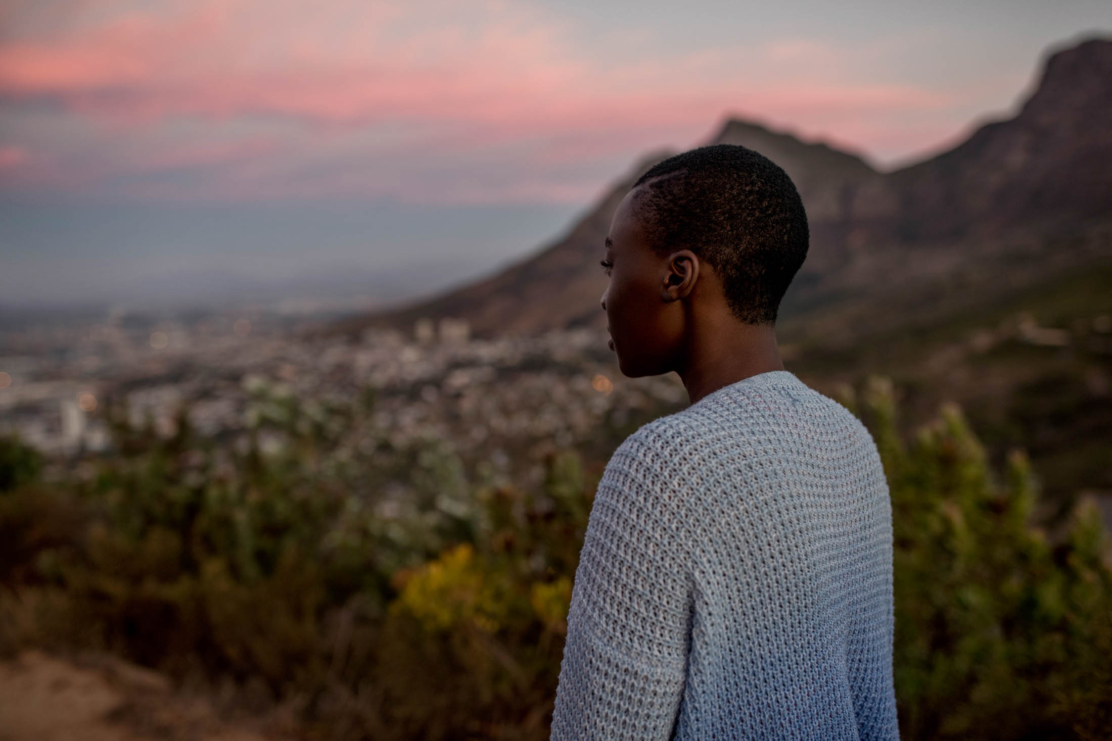 Eine Person blickt bei Sonnenuntergang auf eine Stadtlandschaft mit Bergen im Hintergrund. Der Himmel zeigt Rosa- und Blautöne. © Fotografie Tomas Rodriguez