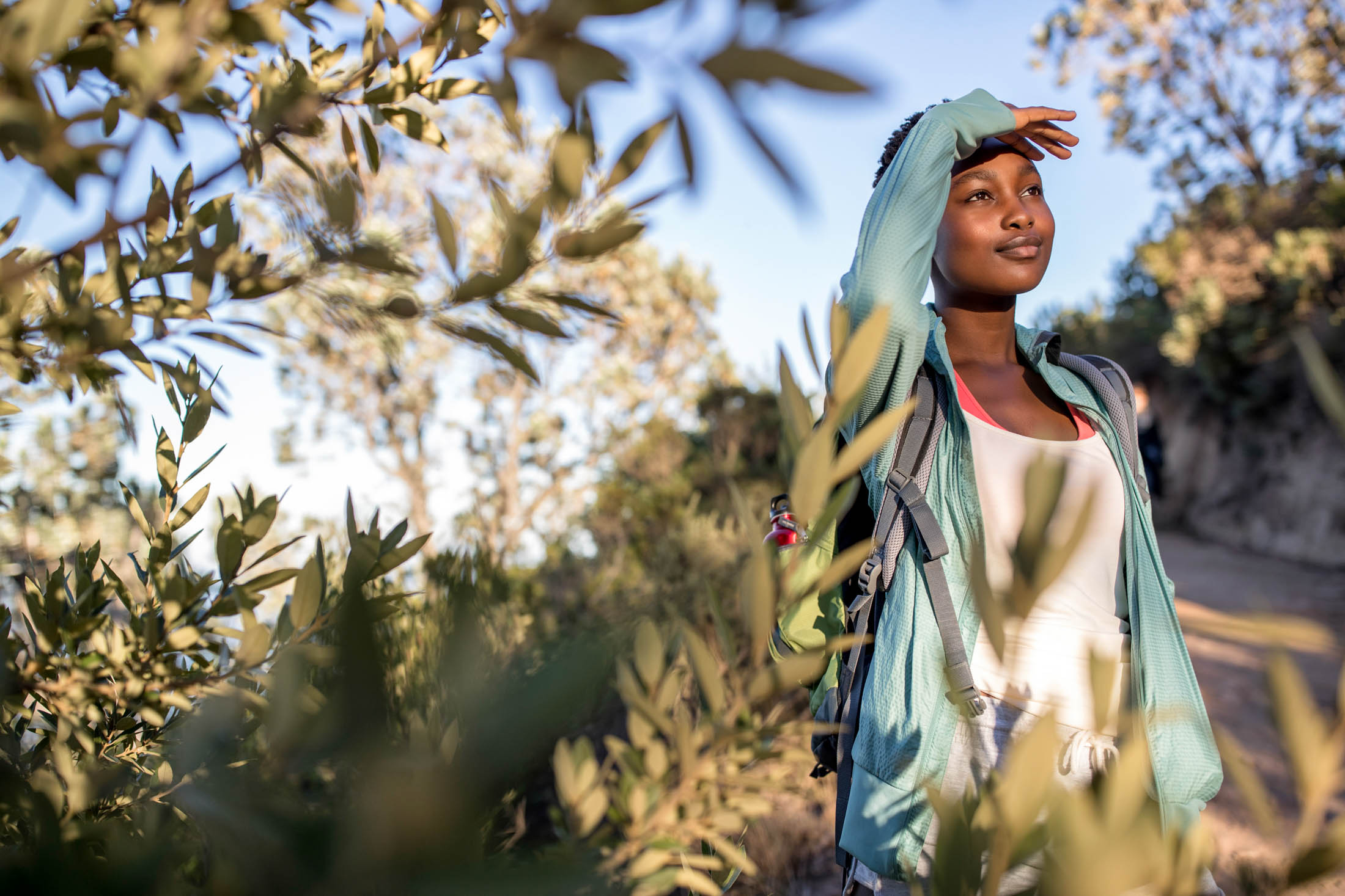 Eine junge Frau wandert an einem sonnigen Tag in der Natur. Sie trägt einen Rucksack und schützt ihre Augen vor der Sonne, umgeben von üppigem Grün. © Fotografie Tomas Rodriguez