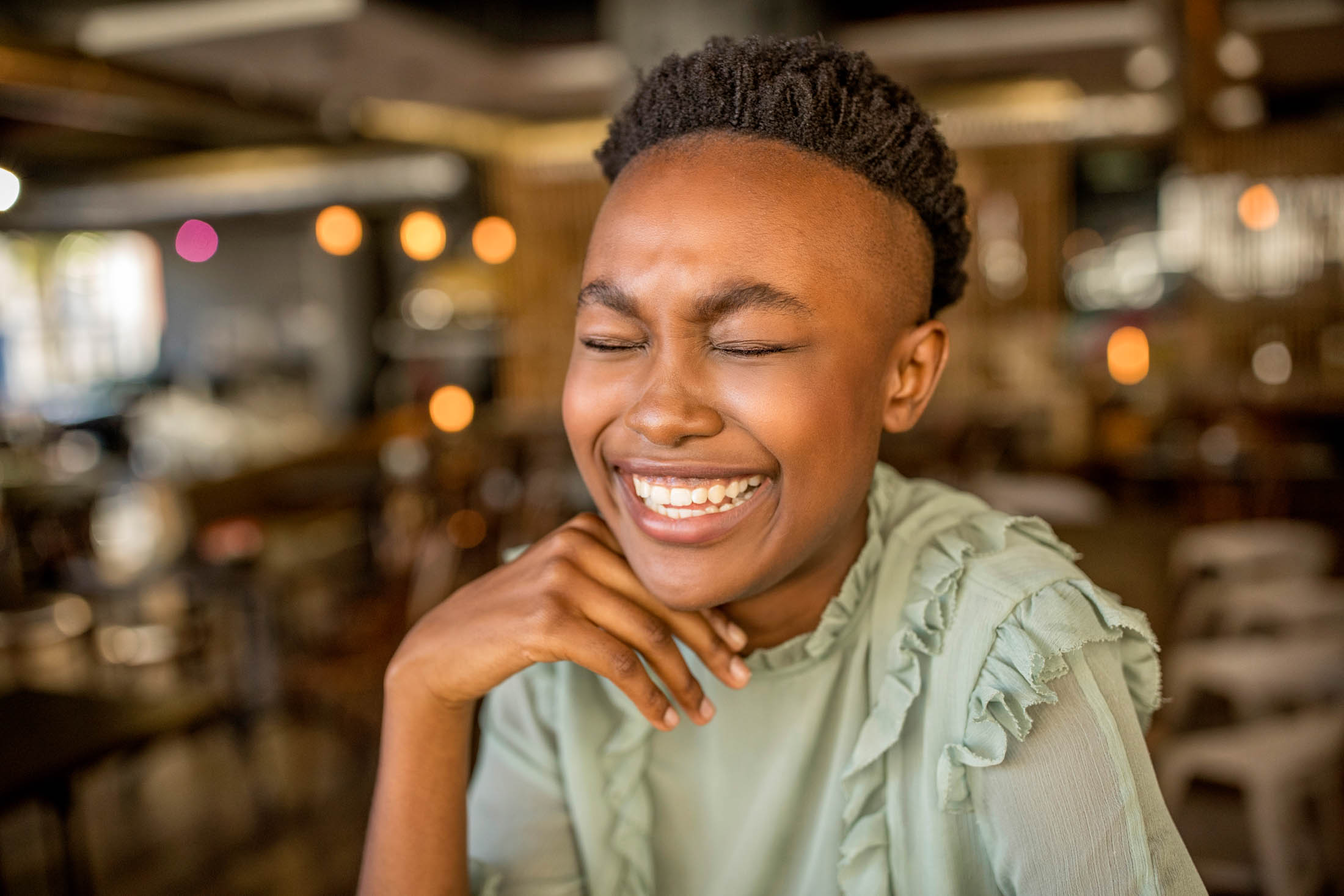Eine fröhliche junge schwarze Frau lacht mit geschlossenen Augen und der Hand am Kinn in einem belebten Café. Ihr Gesichtsausdruck zeugt von echtem Glück. © Fotografie Tomas Rodriguez