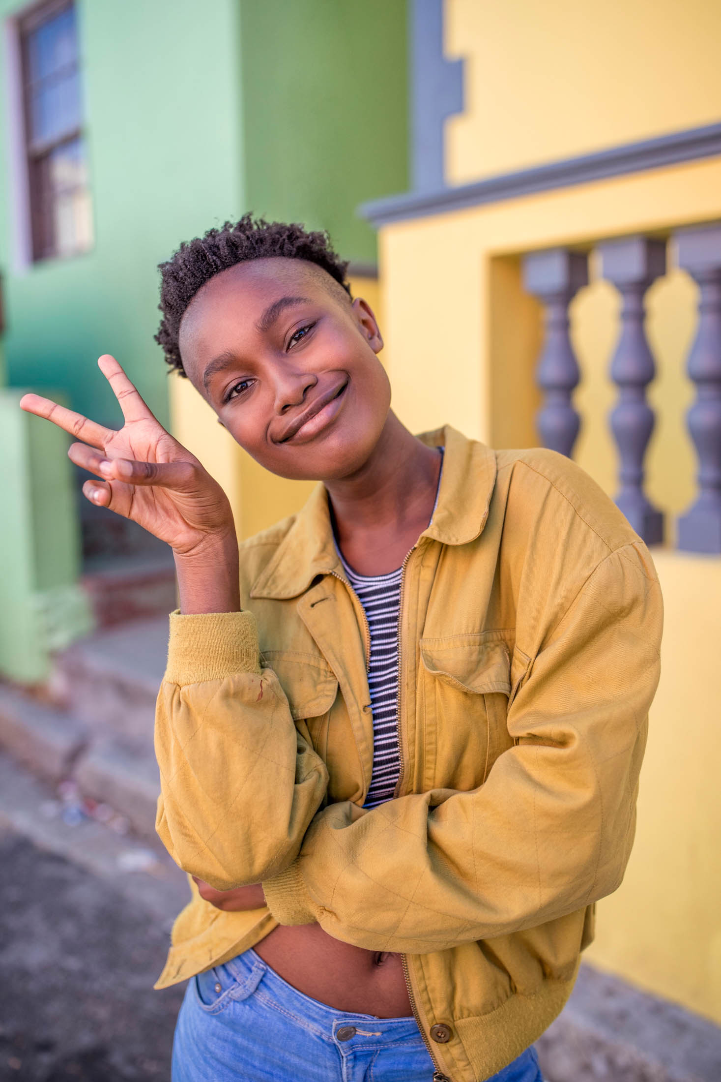 Eine junge Frau mit kurzen Haaren lächelt in die Kamera und macht mit einer Hand ein Peace-Zeichen. Sie trägt ein gestreiftes Hemd und eine gelbe Jacke. Sie steht vor einem farbenfrohen städtischen Hintergrund. © Fotografie Tomas Rodriguez