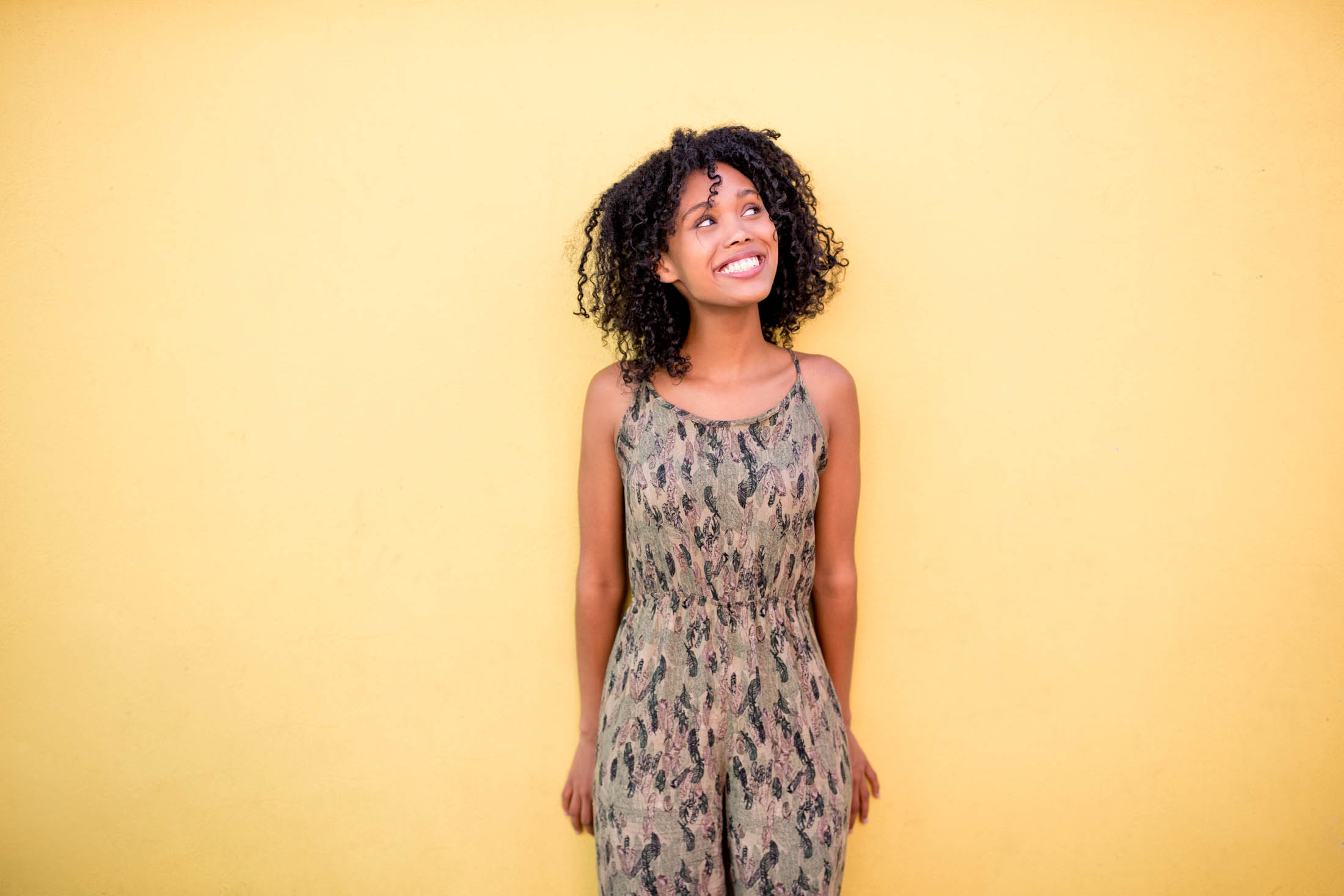 Eine fröhliche junge Frau mit lockigem Haar und einem Tarnkleid, die vor einem leuchtend gelben Hintergrund steht, lächelt und nach oben blickt. © Fotografie Tomas Rodriguez