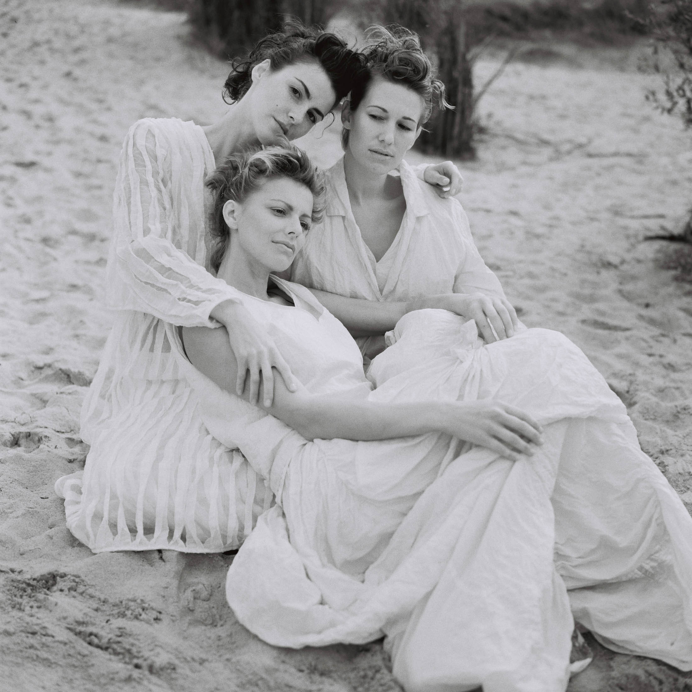 Drei Frauen in fließenden weißen Kleidern sitzen eng beieinander an einem Sandstrand und zeigen heitere und nachdenkliche Ausdrücke. Ihre ineinander verschlungenen Posen vermitteln ein Gefühl von Intimität und Unterstützung. © Fotografie Tomas Rodriguez