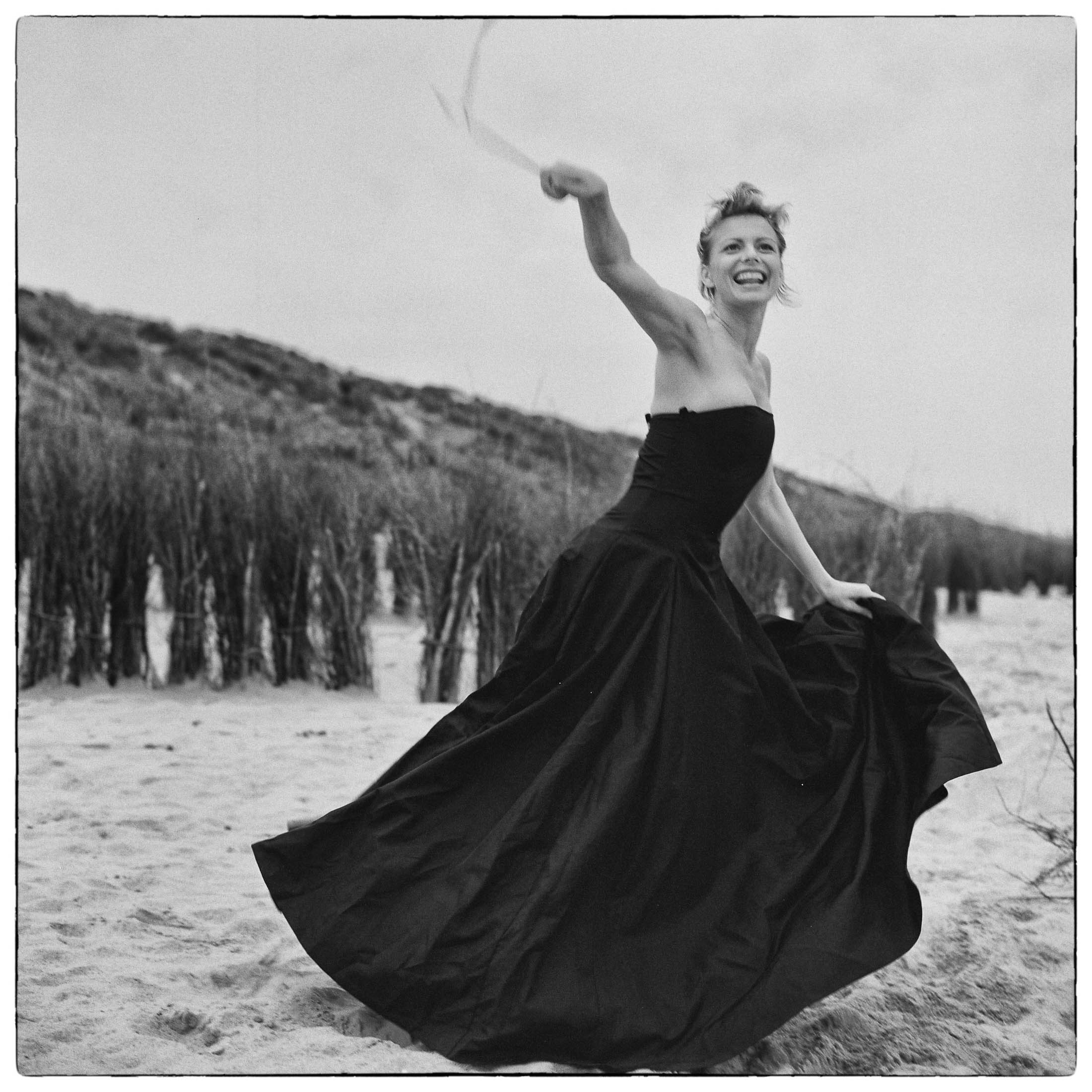 Eine fröhliche Frau in einem fließenden schwarzen Kleid wirbelt auf einem Sandstrand mit Dünen und spärlicher Vegetation im Hintergrund herum. Ihren Arm hat sie erhoben und hält ein dünnes Band. Ihr Gesichtsausdruck ist lebhaft und heiter. © Fotografie Tomas Rodriguez