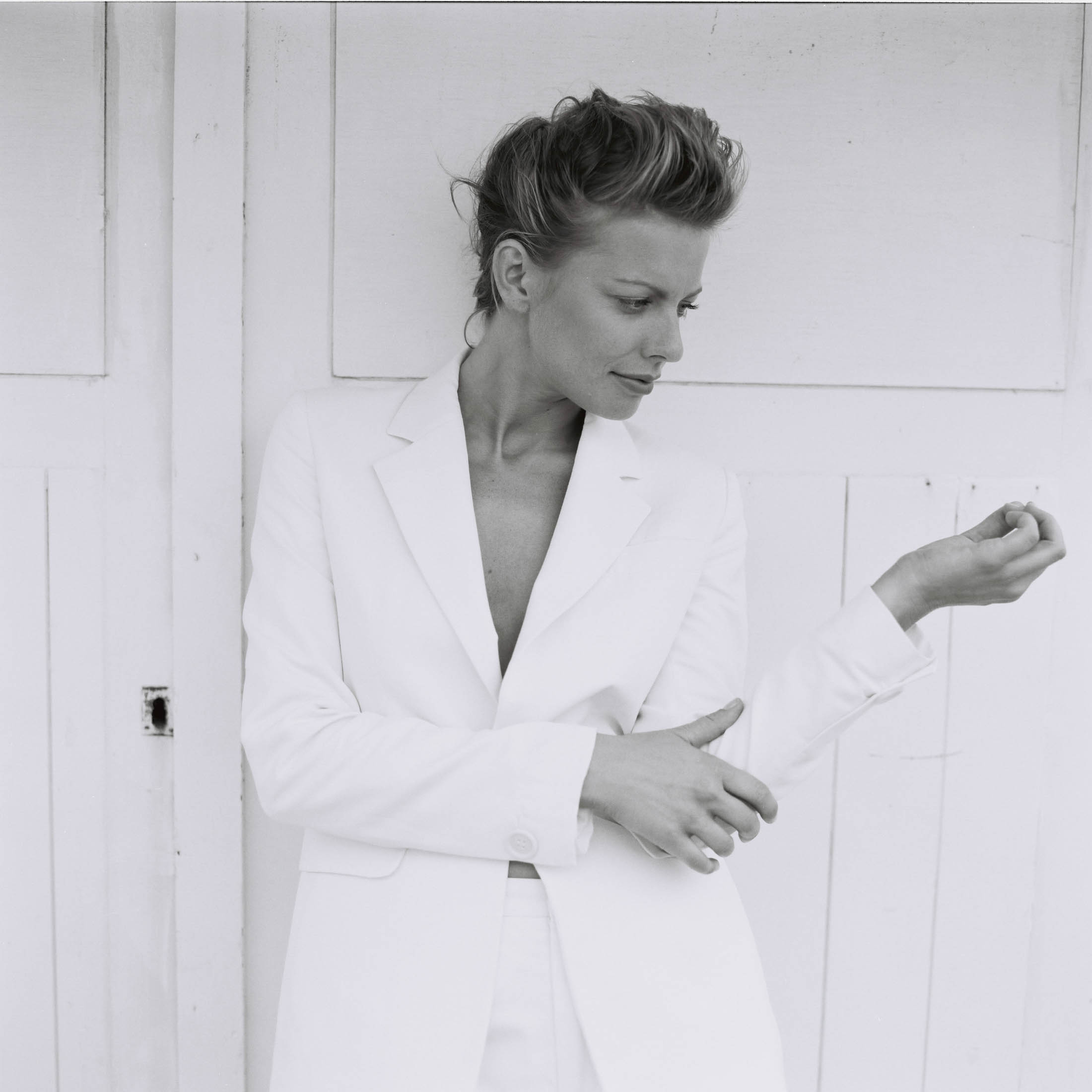 Ein Schwarzweißporträt einer Frau in einem weißen Blazer, die von der Kamera wegblickt und ihren Ärmel vor einem weißen Holzhintergrund zurechtrückt. © Fotografie Tomas Rodriguez