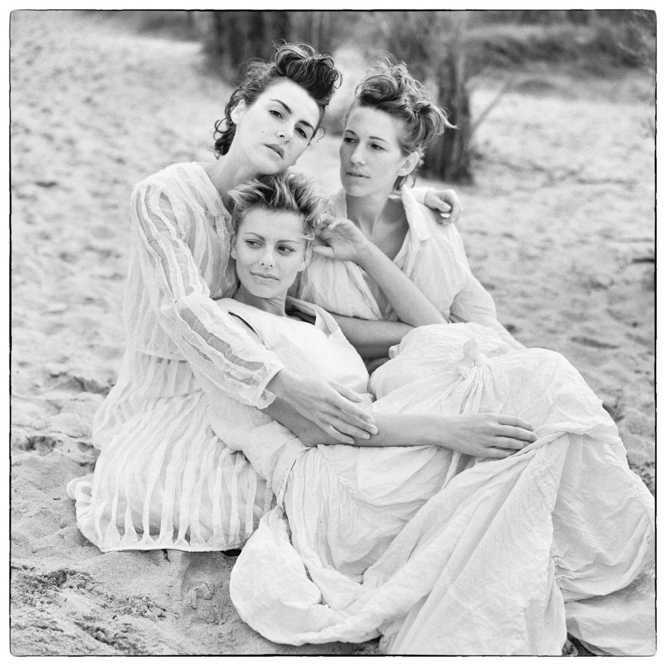 Drei Frauen in fließenden weißen Kleidern sitzen eng beieinander an einem Sandstrand und posieren nachdenklich für ein Schwarzweißfoto. © Fotografie Tomas Rodriguez