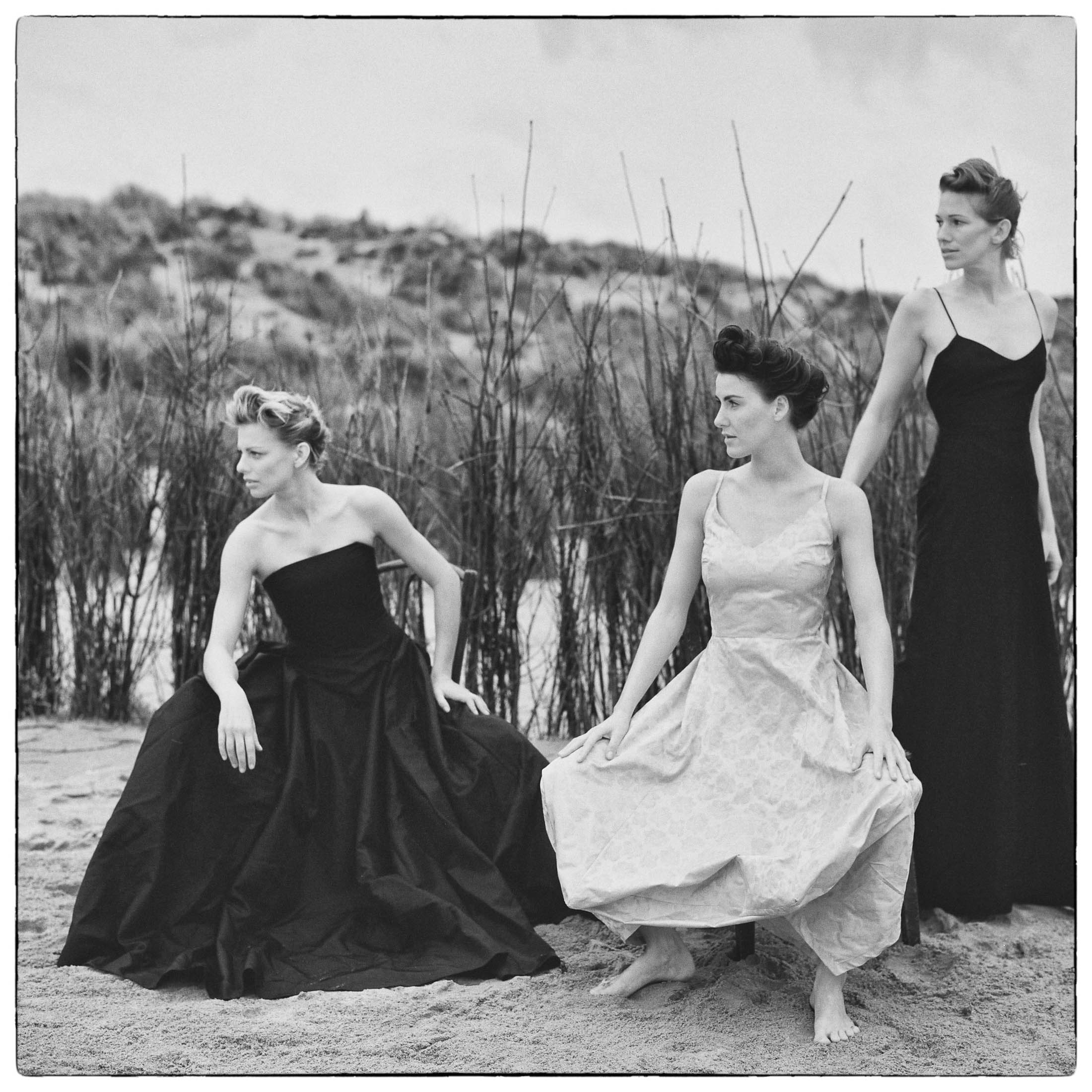 Drei Frauen in eleganten Kleidern stehen barfuß auf einer Sandfläche mit hohem Gras und wirken nachdenklich unter einem bewölkten Himmel. Die Kulisse deutet auf ein Vintage- oder zeitloses Thema hin. © Fotografie Tomas Rodriguez
