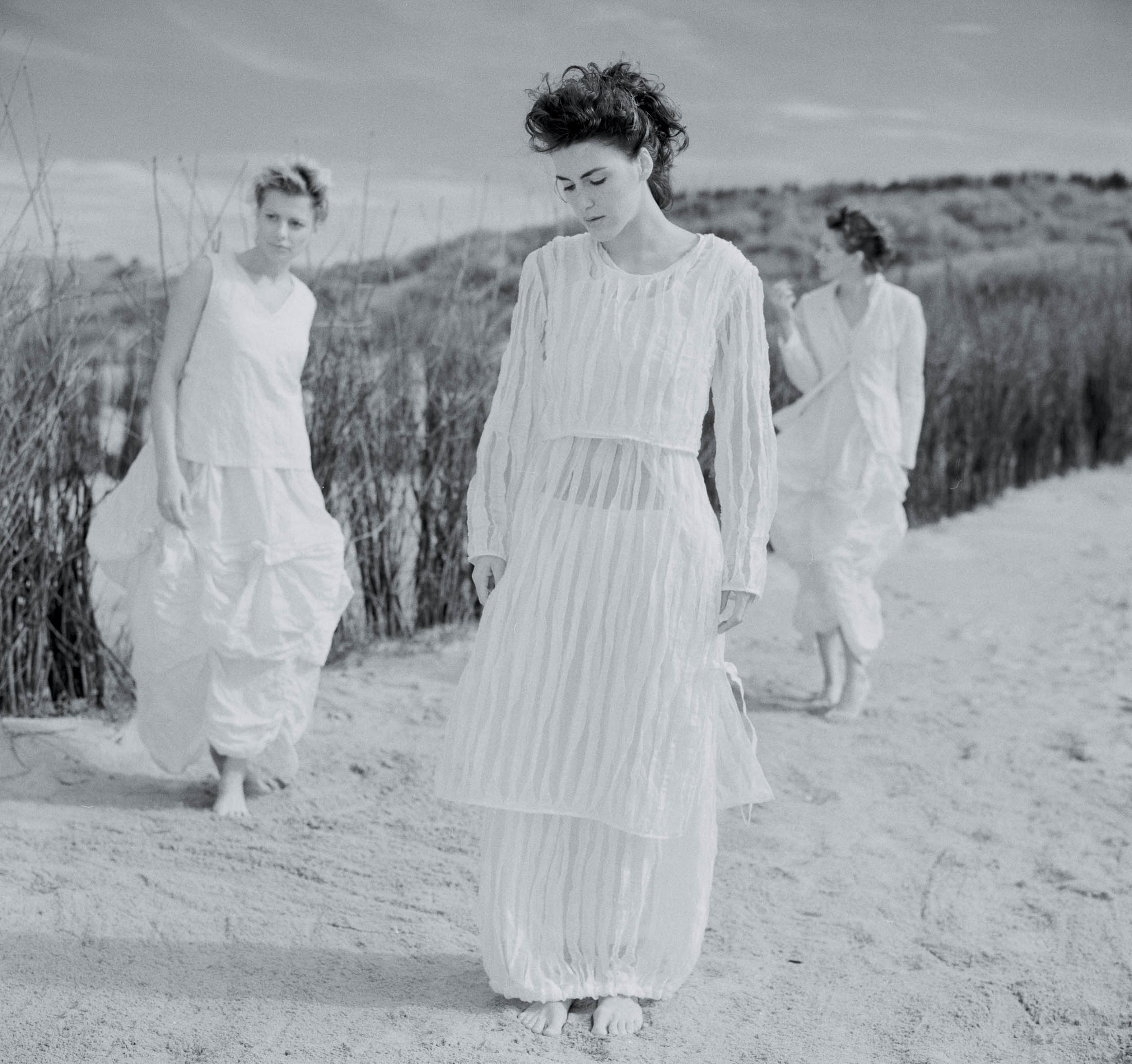 Drei Frauen in weißen Kleidern gehen an einem Sandstrand spazieren, eine im Vordergrund blickt nach unten, die anderen im Hintergrund zwischen hohem Gras. Die Kulisse ruft eine heitere, zeitlose Atmosphäre hervor. © Fotografie Tomas Rodriguez