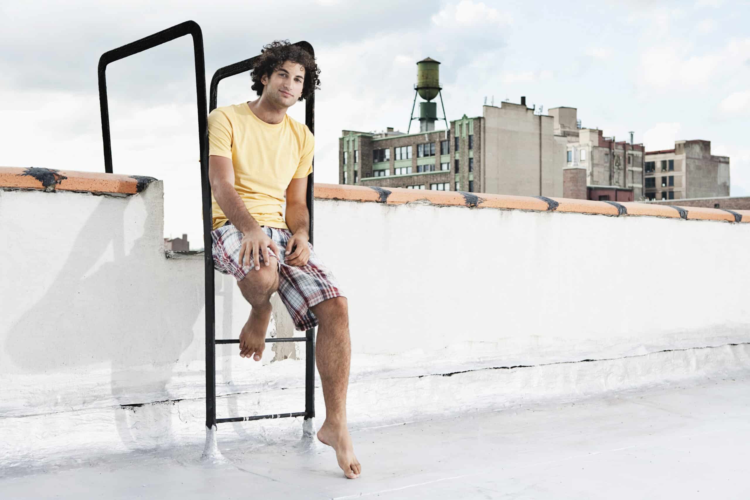 Eine Person mit lockigem Haar, einem gelben T-Shirt und karierten Shorts trägt, sitzt auf einer Leiter vor einer weißen Wand auf einem Dach. Ein Fuß steht auf der Leiter, der andere baumelt. Im Hintergrund ist eine urbane Skyline mit alten Gebäuden und einem Wasserturm zu sehen. © Fotografie Tomas Rodriguez