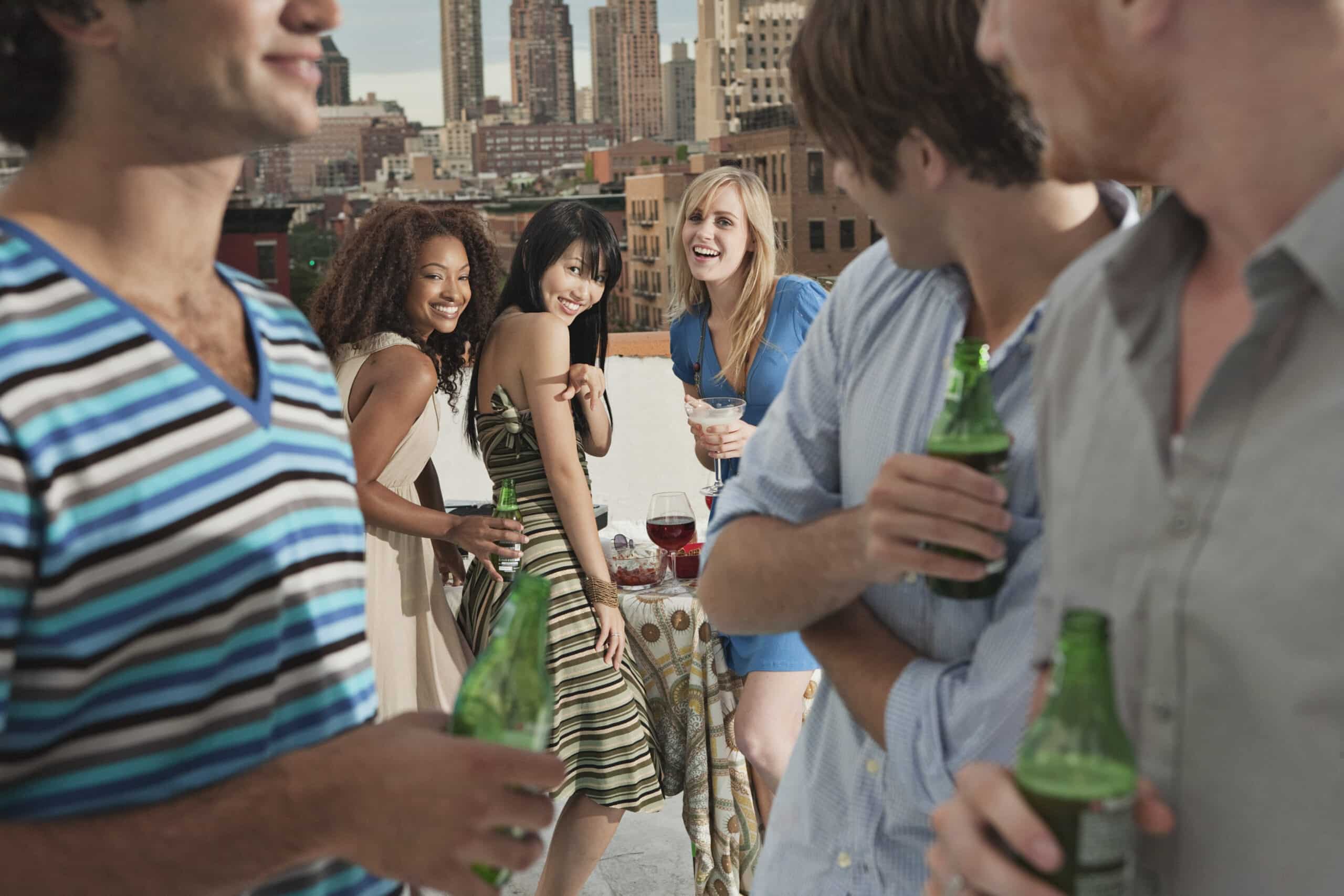 Eine Gruppe von Freunden trifft sich auf einem Dach. Drei Frauen lächeln und posieren für ein Foto an einem Tisch mit Getränken. Zwei Männer im Vordergrund halten Bierflaschen und schauen in ihre Richtung. Im Hintergrund sind Stadtgebäude zu sehen. © Fotografie Tomas Rodriguez