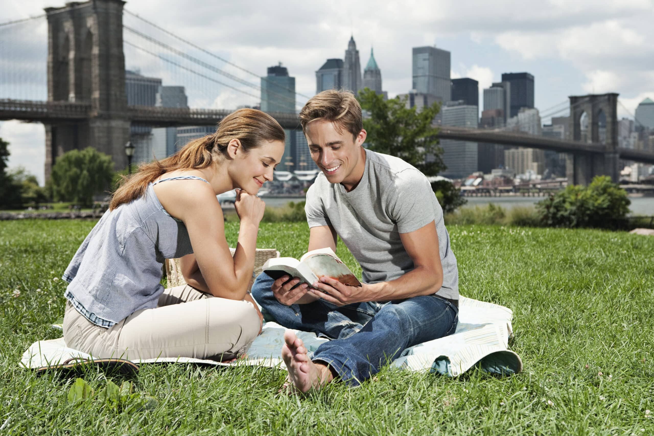 Ein Mann und eine Frau sitzen auf einer Decke in einem Park und lesen gemeinsam Bücher. Im Hintergrund sind die Brooklyn Bridge und die Skyline einer Stadt zu sehen. Die Frau lächelt und stützt ihr Kinn auf ihre Hand, während der Mann ein Buch hält und laut vorzulesen scheint. © Fotografie Tomas Rodriguez