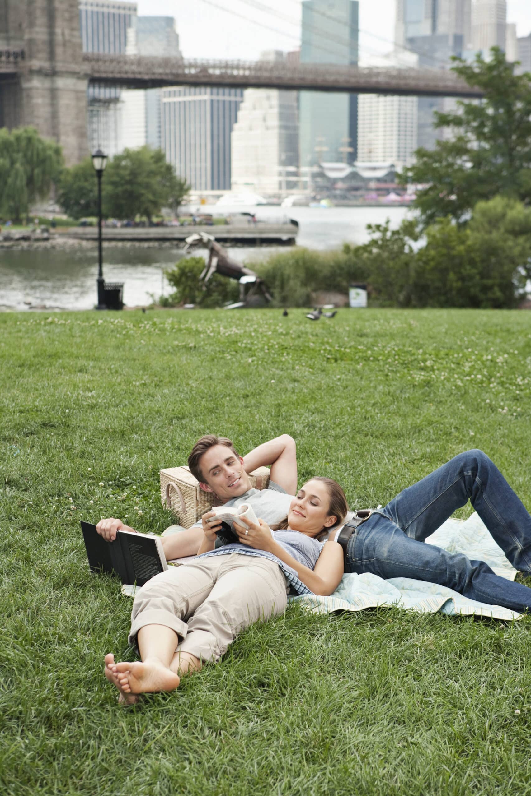 Ein Paar entspannt sich auf einer Picknickdecke in einem Park. Eine Person liegt auf dem Rücken und liest ein Buch, während die andere sich an sie lehnt und in ihr Handy vertieft ist. Im Hintergrund sind die Skyline der Stadt und die Brooklyn Bridge sowie üppige grüne Bäume und Gras zu sehen. © Fotografie Tomas Rodriguez
