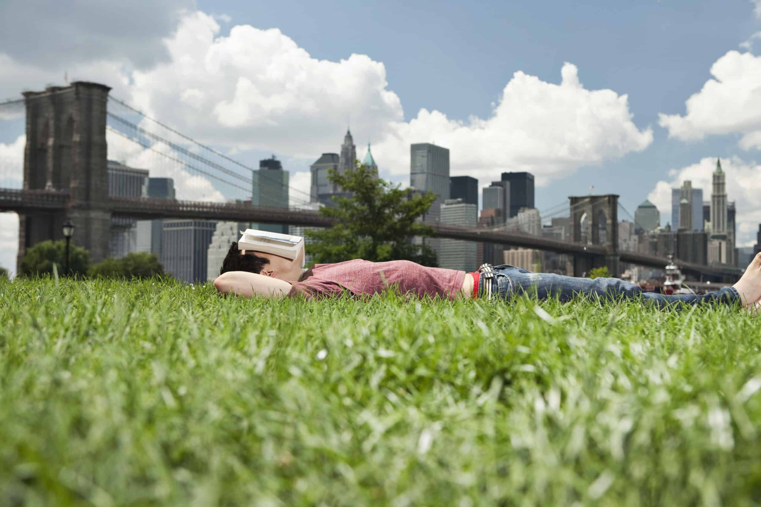 Eine Person liegt auf üppigem grünem Gras und hat ein geschlossenes Buch vor dem Gesicht. Sie entspannt sich unter einem teilweise bewölkten Himmel. Im Hintergrund sind eine große Hängebrücke und die Skyline einer Stadt mit hohen Gebäuden zu sehen. © Fotografie Tomas Rodriguez
