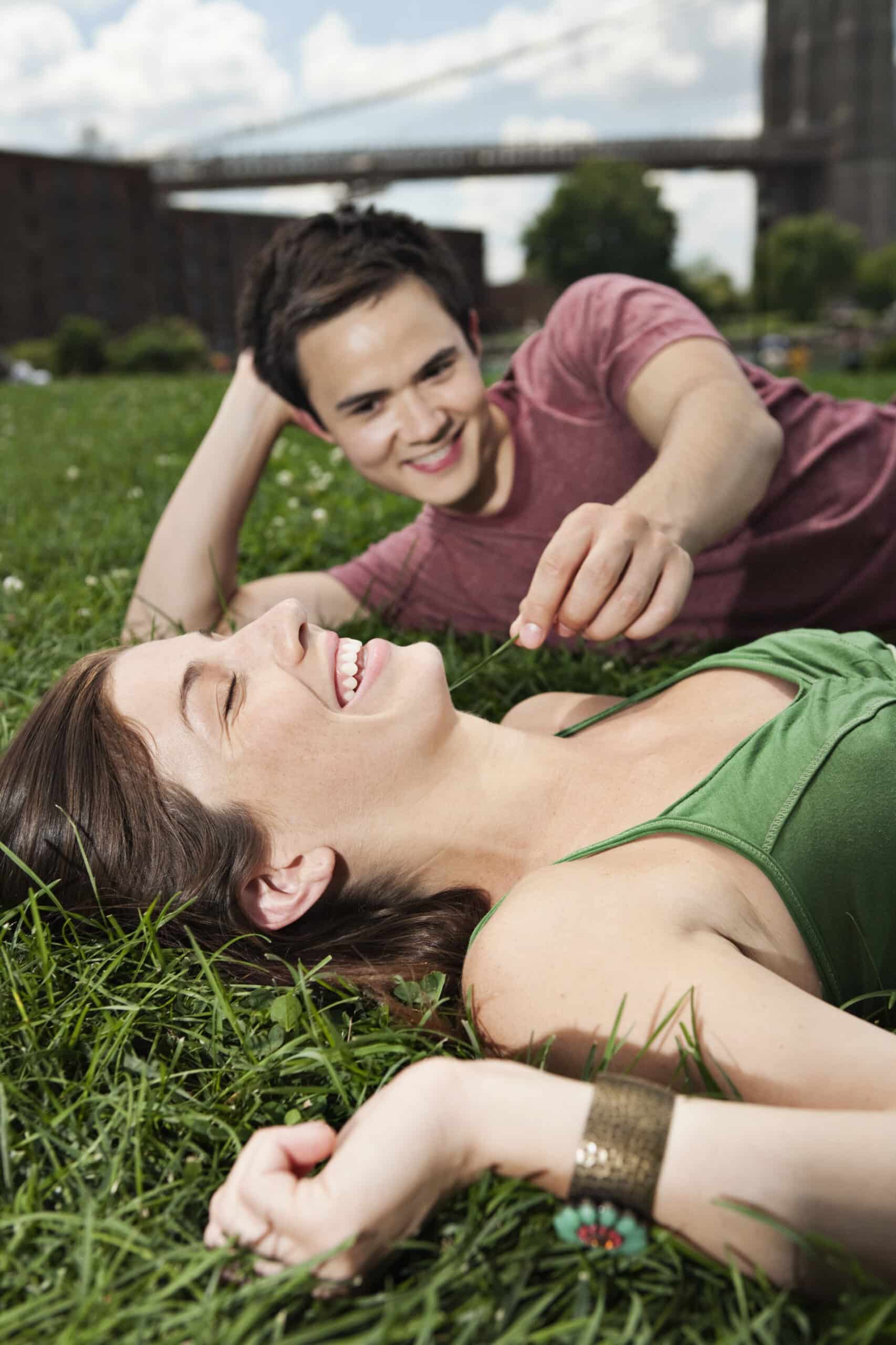 Ein Mann und eine Frau liegen an einem sonnigen Tag im Gras eines Parks. Die Frau, die ein grünes Oberteil trägt, lacht, während der Mann, der ein rotes Hemd trägt, spielerisch nach ihr greift. Im Hintergrund sind Bäume und eine Brücke zu sehen. © Fotografie Tomas Rodriguez