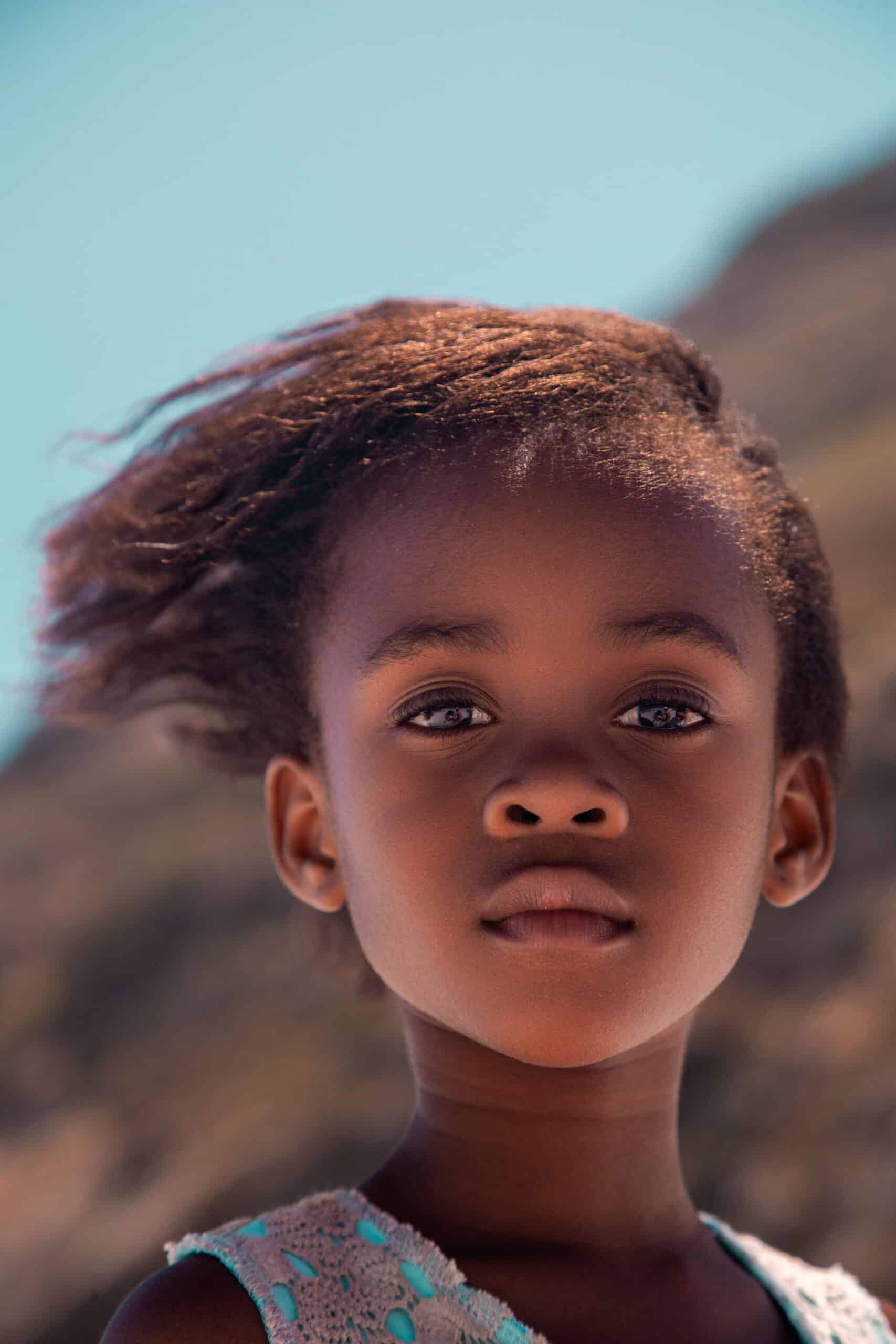 Ein junges Mädchen mit nachdenklichem Gesichtsausdruck und im Wind wehendem Haar steht im Freien vor einem unscharfen natürlichen Hintergrund. Sie hat große Augen und trägt ein hellblaues Kleid. © Fotografie Tomas Rodriguez