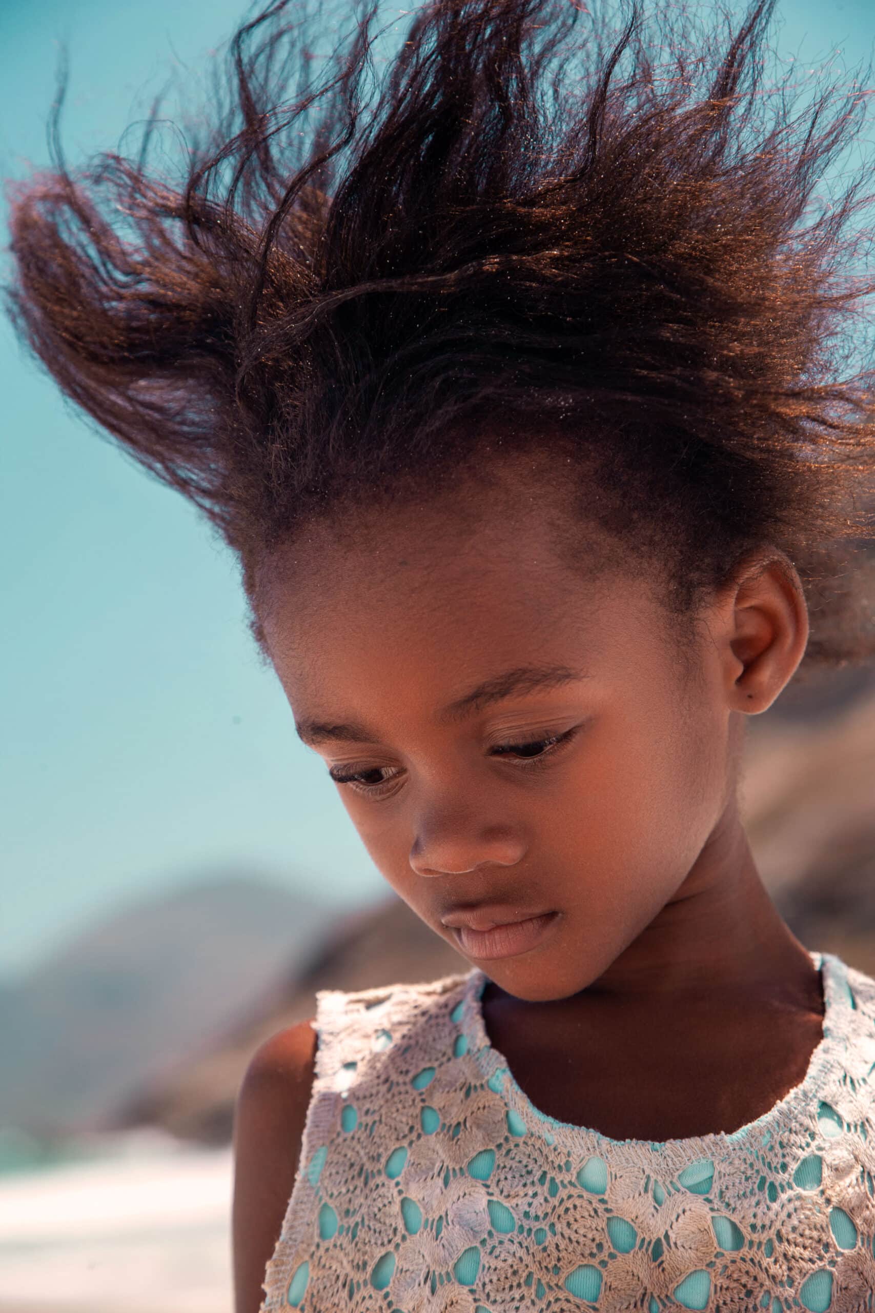 Ein junges Mädchen mit im Wind wehendem Haar, das mit nachdenklichem Gesichtsausdruck vor der Kulisse eines Strandes nach unten blickt. © Fotografie Tomas Rodriguez