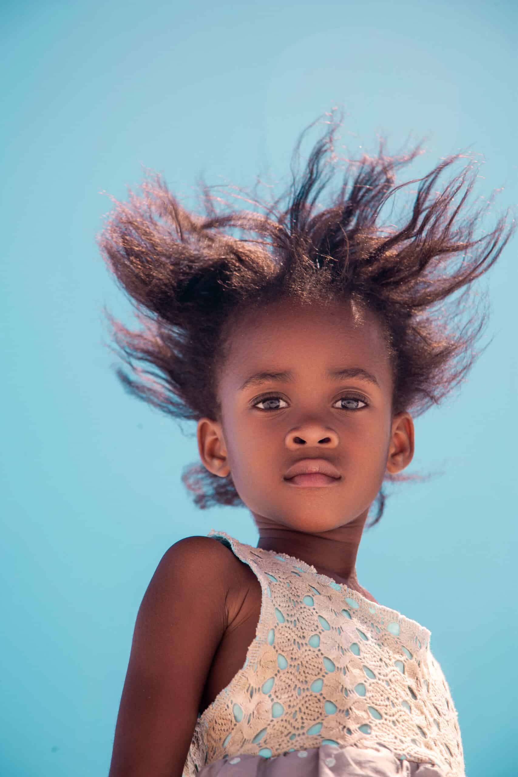 Ein junges Mädchen mit vom Wind zerzaustem Haar steht vor einem klaren blauen Himmel, trägt ein weißes Spitzenkleid und blickt nachdenklich in die Ferne. © Fotografie Tomas Rodriguez