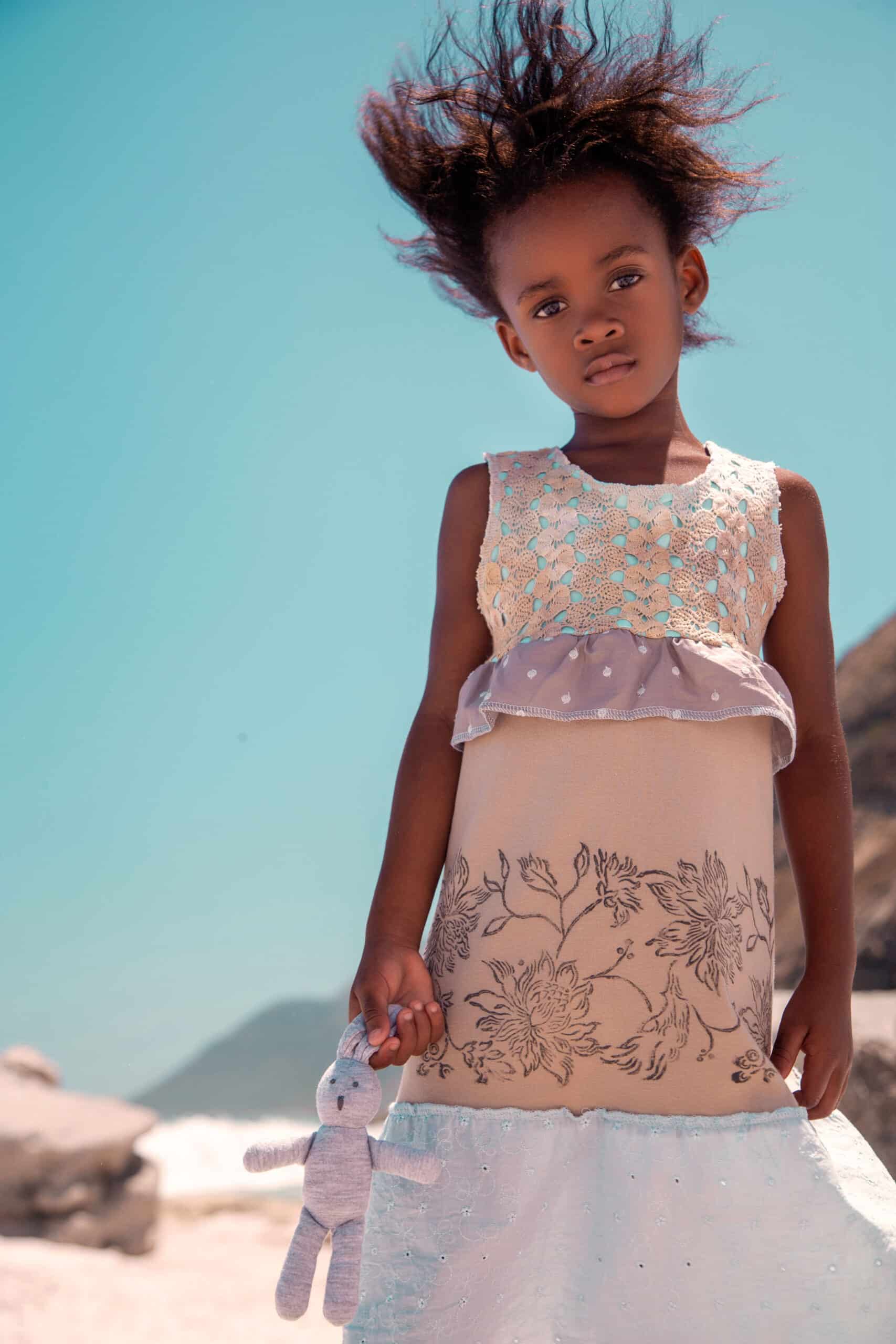 Ein junges Mädchen steht mit einem Spielzeug in der Hand an einem Strand, wobei ihr Haar vom Wind nach oben geweht wird. Sie trägt ein detailreiches, elegantes Kleid mit Blumenmuster vor dem Hintergrund eines blauen Himmels und einer felsigen Landschaft. © Fotografie Tomas Rodriguez