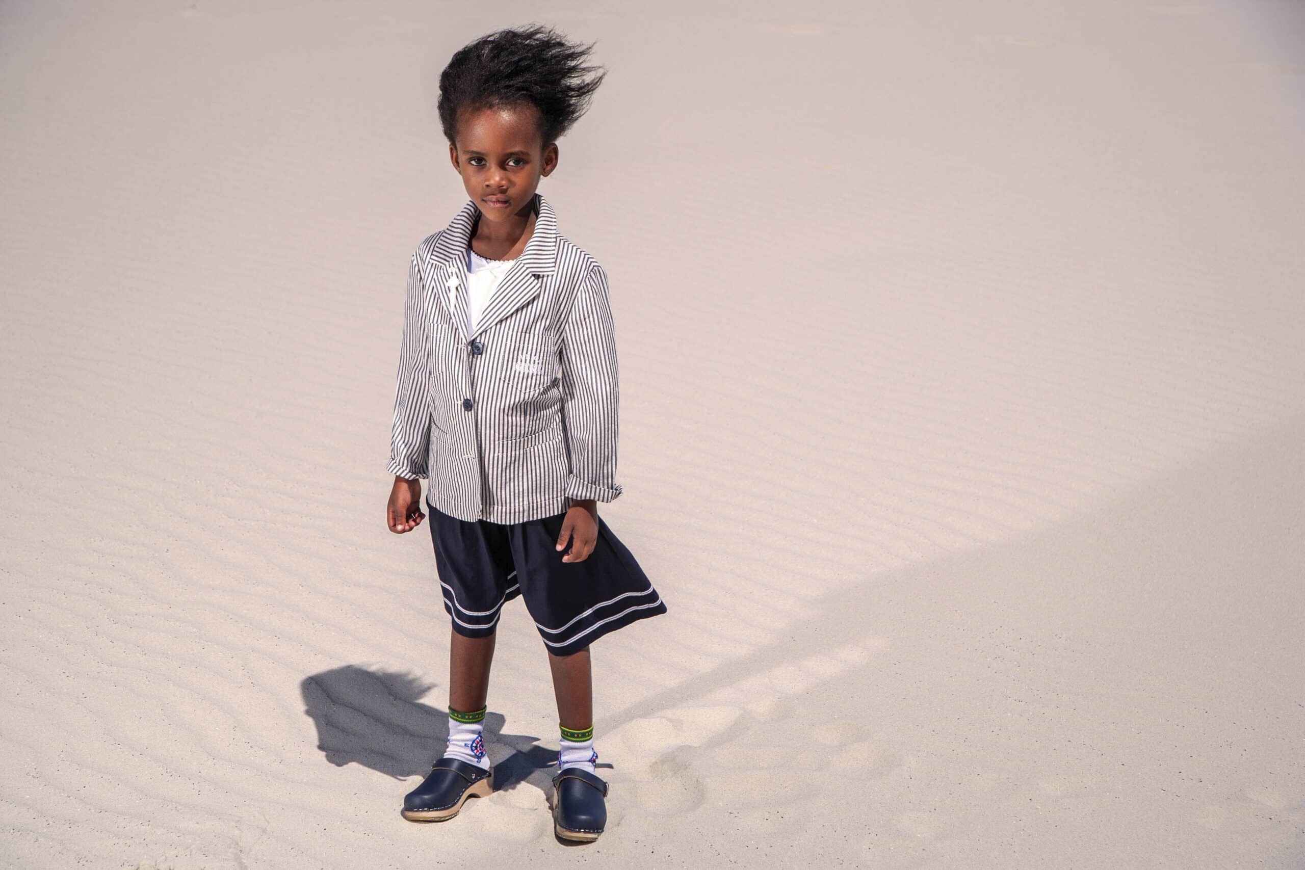 Ein kleines Kind steht auf einem Sandboden. Es trägt einen übergroßen gestreiften Blazer und Shorts und hat vom Wind zerzauste Haare. © Fotografie Tomas Rodriguez