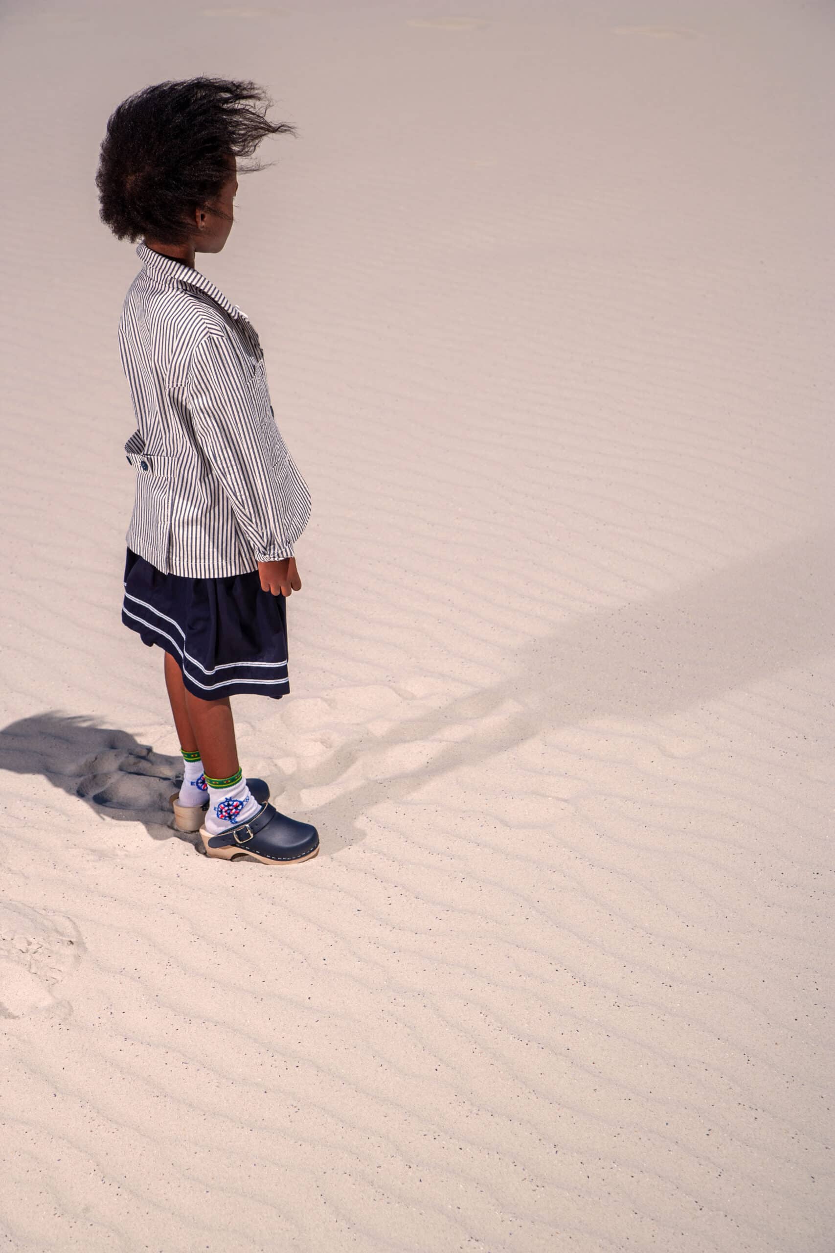 Ein kleines Kind mit vollem Haar läuft auf einem Sandboden und hinterlässt Fußabdrücke. Das Kind trägt eine gestreifte Jacke und einen dunklen Rock und hellblaue Schuhe. © Fotografie Tomas Rodriguez