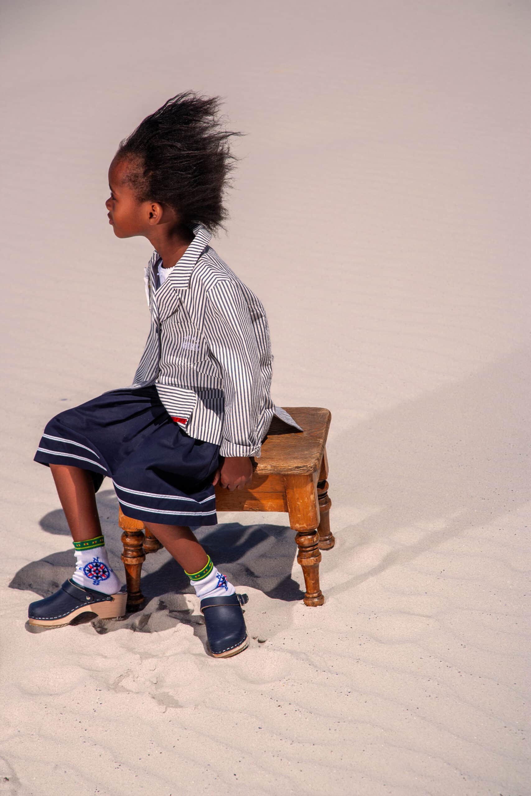 Ein kleines Kind mit einer üppigen Frisur sitzt auf einem alten Holzhocker auf sandigem Untergrund und schaut zur Seite. Es trägt ein gestreiftes Hemd, einen marineblauen Rock und verspielte Socken und Schuhe. © Fotografie Tomas Rodriguez