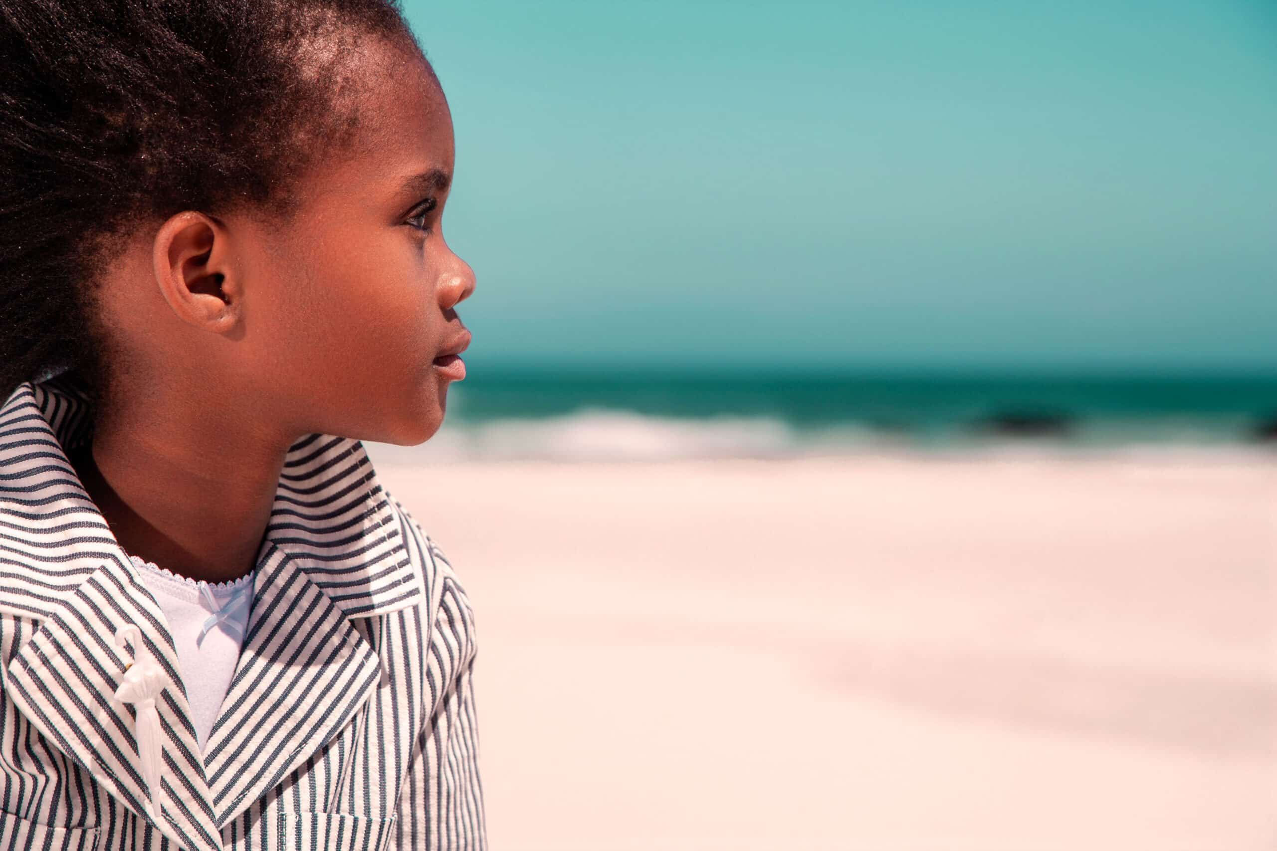 Ein junges Mädchen mit dunklem, zu Zöpfen frisiertem Haar blickt an einem sonnigen Strand aufs Meer. Es trägt eine gestreifte Jacke und hat einen nachdenklichen Gesichtsausdruck. © Fotografie Tomas Rodriguez