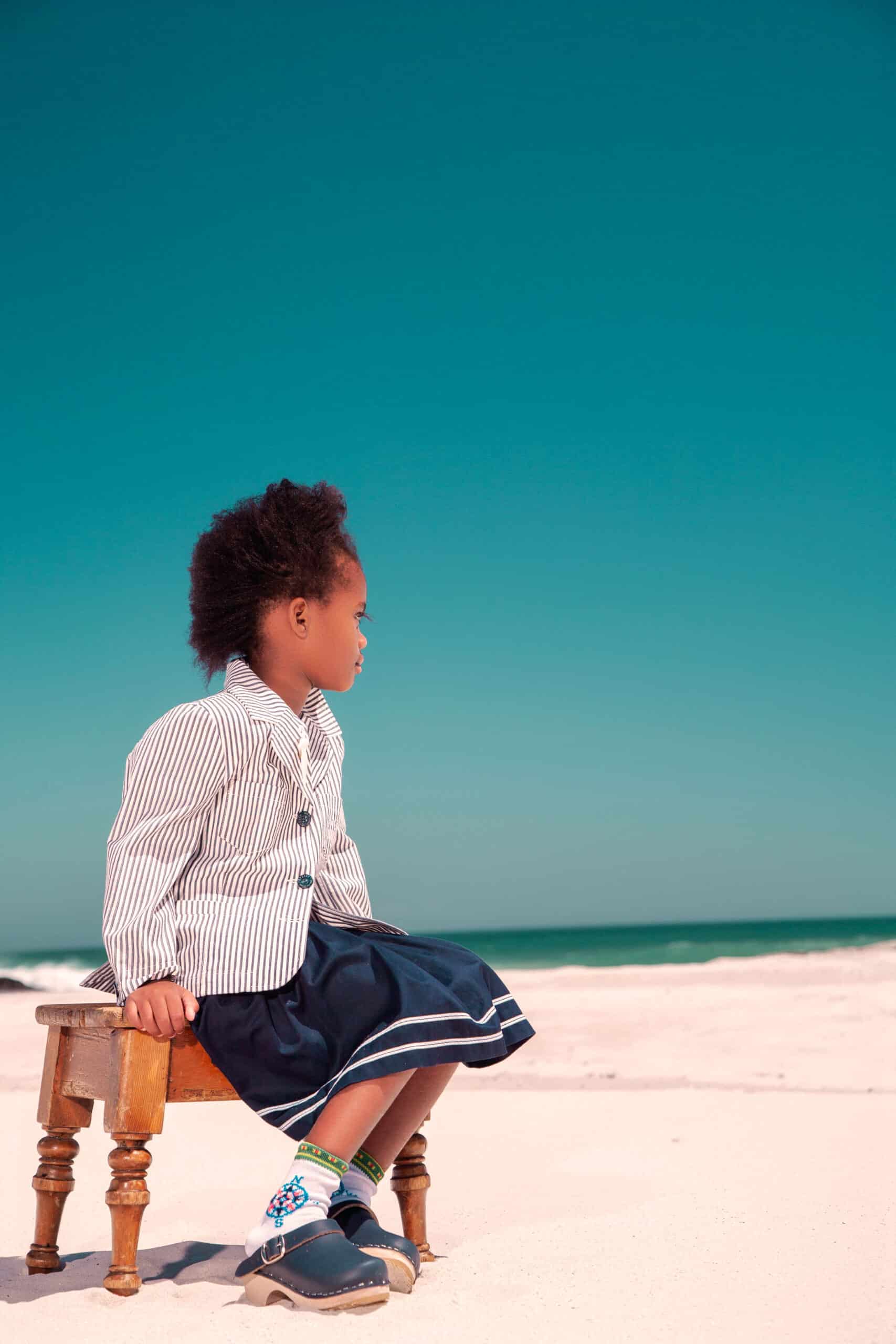 Ein kleines Kind mit dunklem, lockigem Haar, bekleidet mit einem gestreiften Hemd und einem marineblauen Rock, sitzt auf einem verzierten Holzstuhl an einem Sandstrand und blickt unter einem klaren blauen Himmel auf das Meer. © Fotografie Tomas Rodriguez