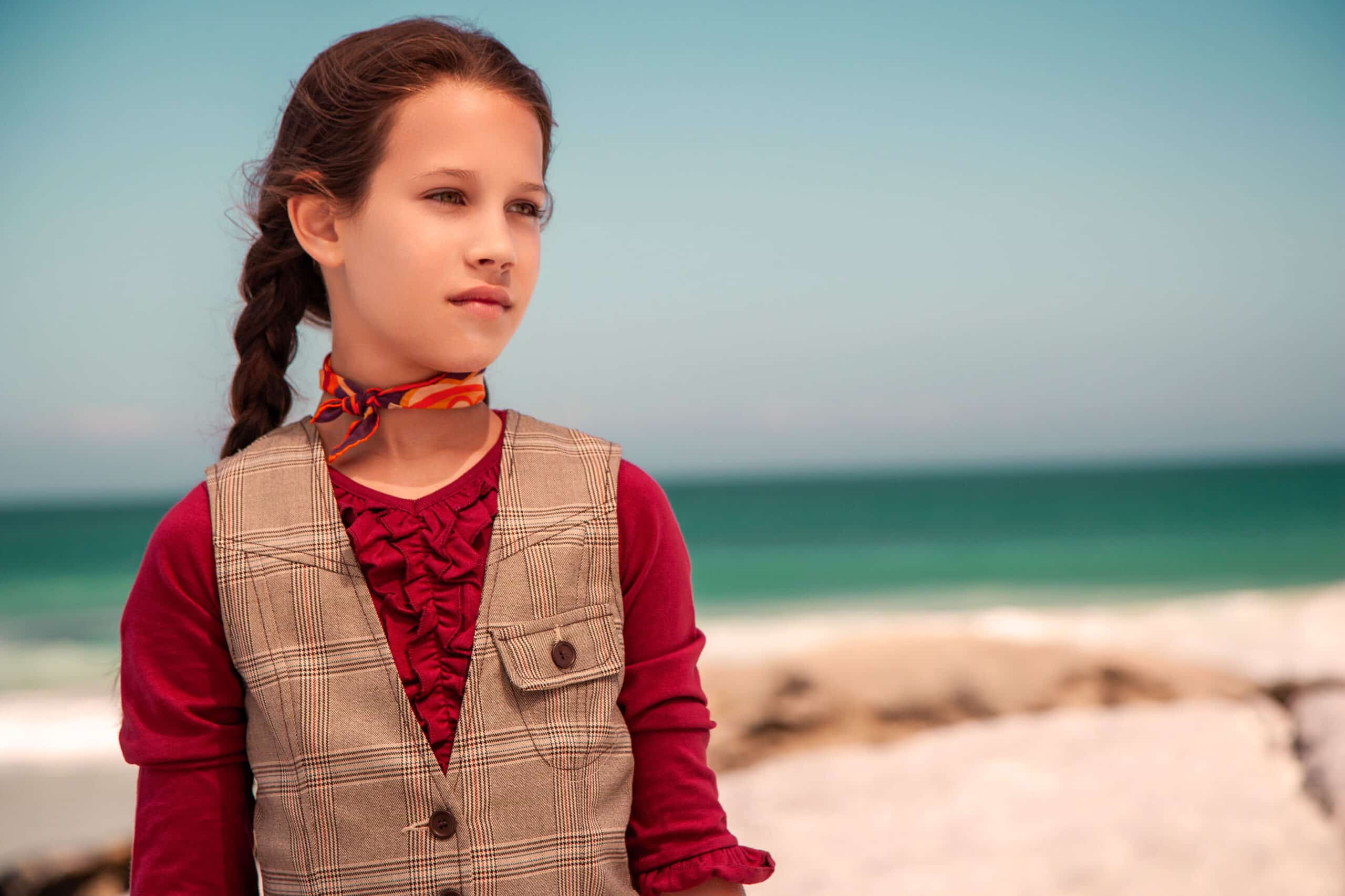 Ein junges Mädchen mit einem seitlichen Zopf, einer Tweedweste und einer roten Bluse steht nachdenklich an einem Strand mit dem Meer im Hintergrund. © Fotografie Tomas Rodriguez