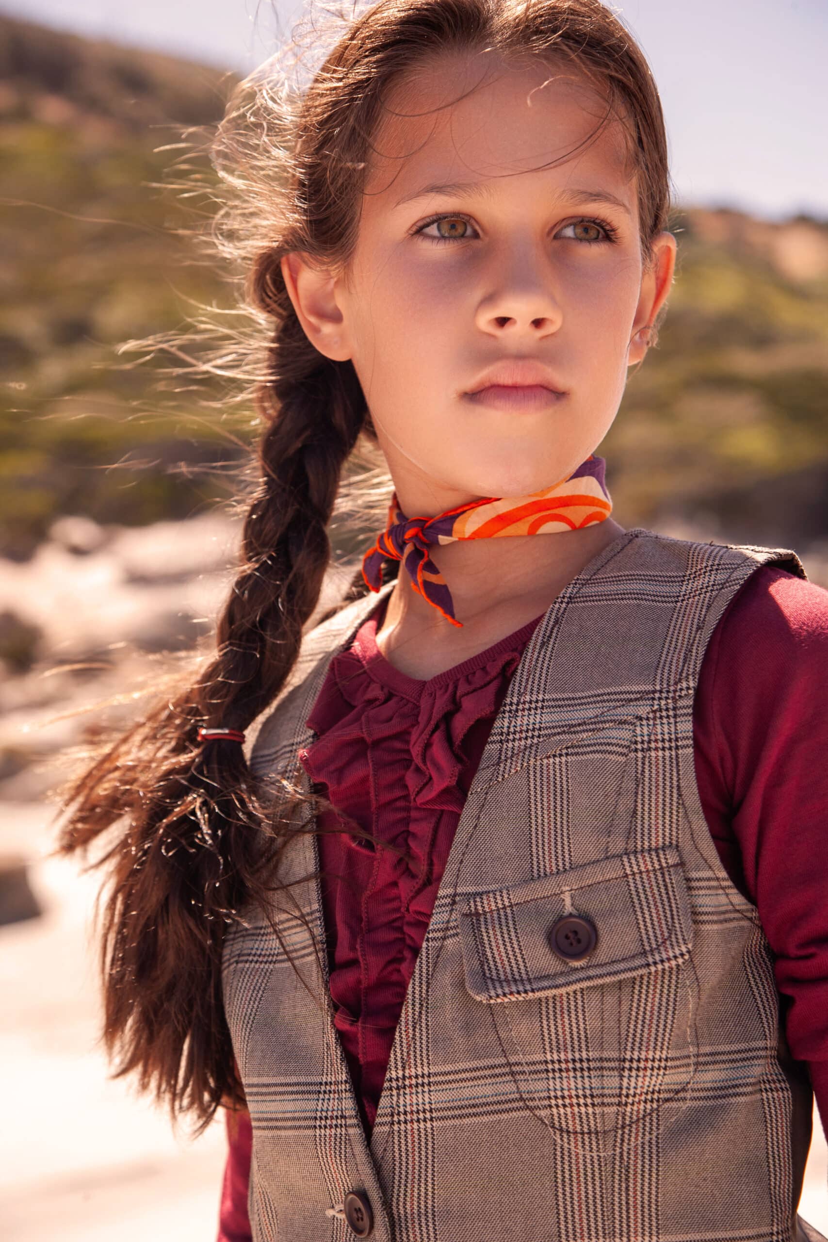 Ein junges Mädchen mit Zopf und nachdenklichem Gesichtsausdruck steht im Freien. Es trägt eine Tweedweste, eine rote Bluse und einen orangefarbenen Schal um den Hals vor einem sandigen und bewachsenen Hintergrund. © Fotografie Tomas Rodriguez