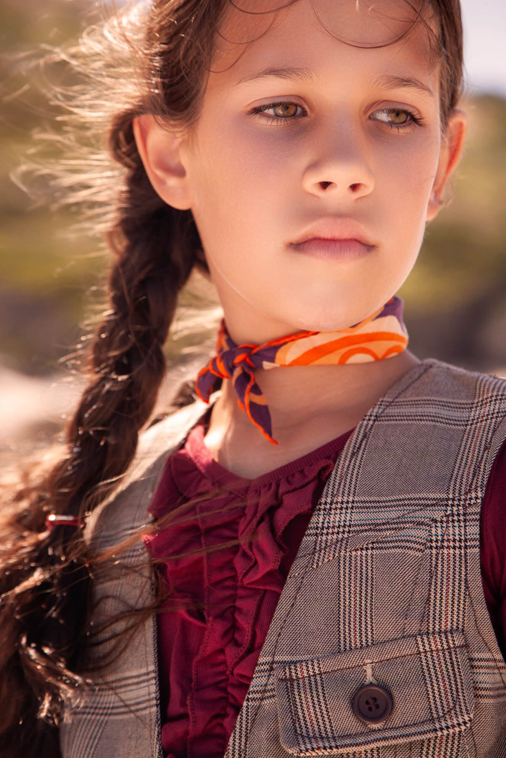 Ein junges Mädchen mit nachdenklichem Gesichtsausdruck, trägt eine Weste und eine kastanienbraune Rüschenbluse, hat einen bunten Schal um den Hals und ihr braunes Haar ist zu einem seitlichen Zopf geflochten. © Fotografie Tomas Rodriguez