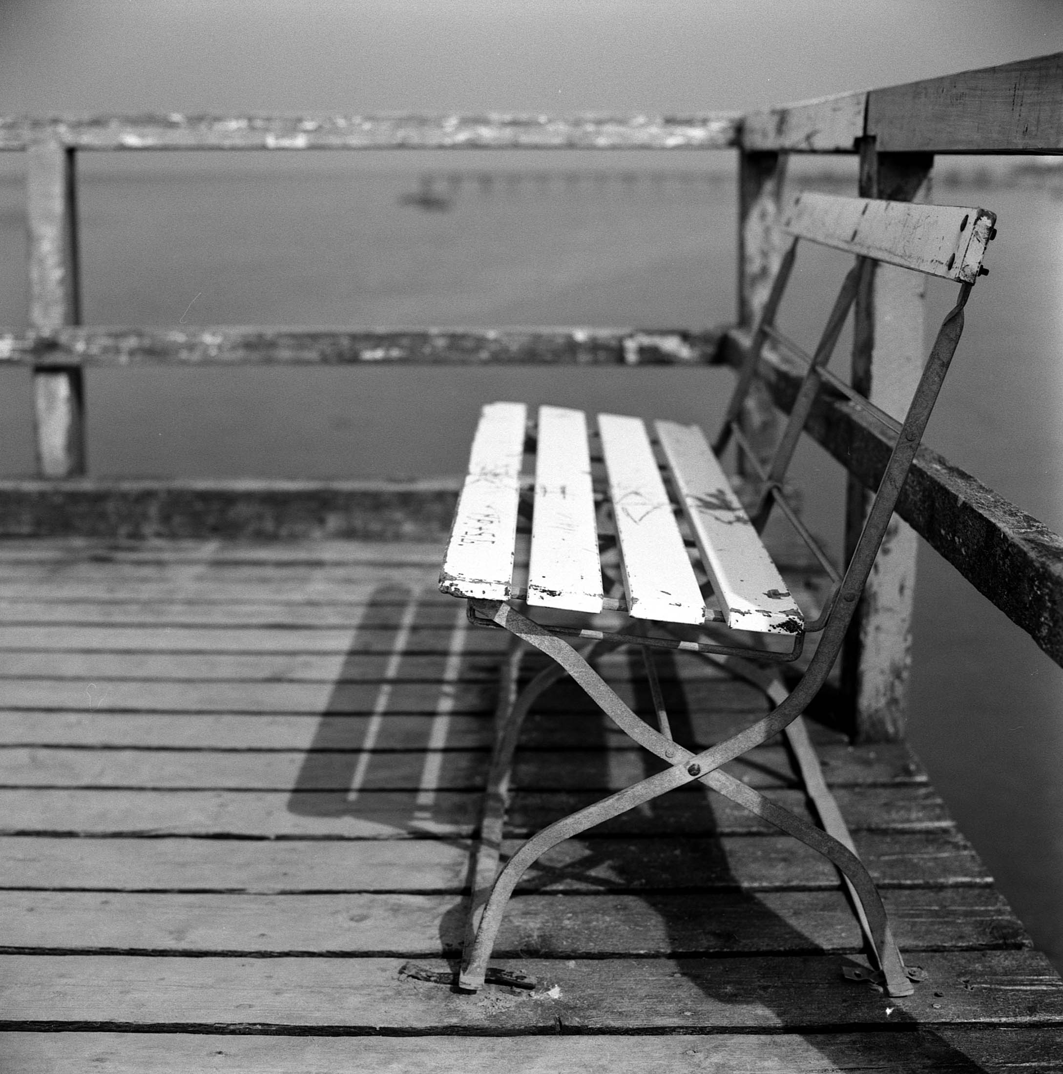 Schwarzweißfoto eines verwitterten weißen Klappstuhls auf einem Holzsteg mit ruhigem Wasser im Hintergrund und einem Holzgeländer, das einen Schatten wirft. © Fotografie Tomas Rodriguez