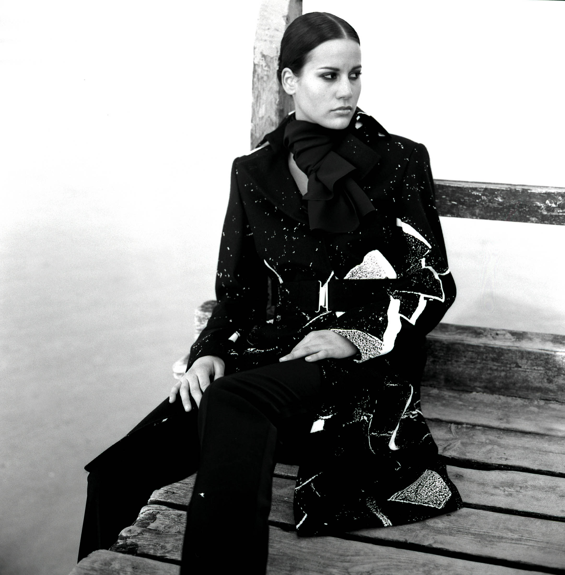 Eine Frau in einem eleganten schwarz-weiß gefleckten Anzug mit einer großen Schleife am Hals sitzt nachdenklich auf einer Holzbank am Wasser. © Fotografie Tomas Rodriguez