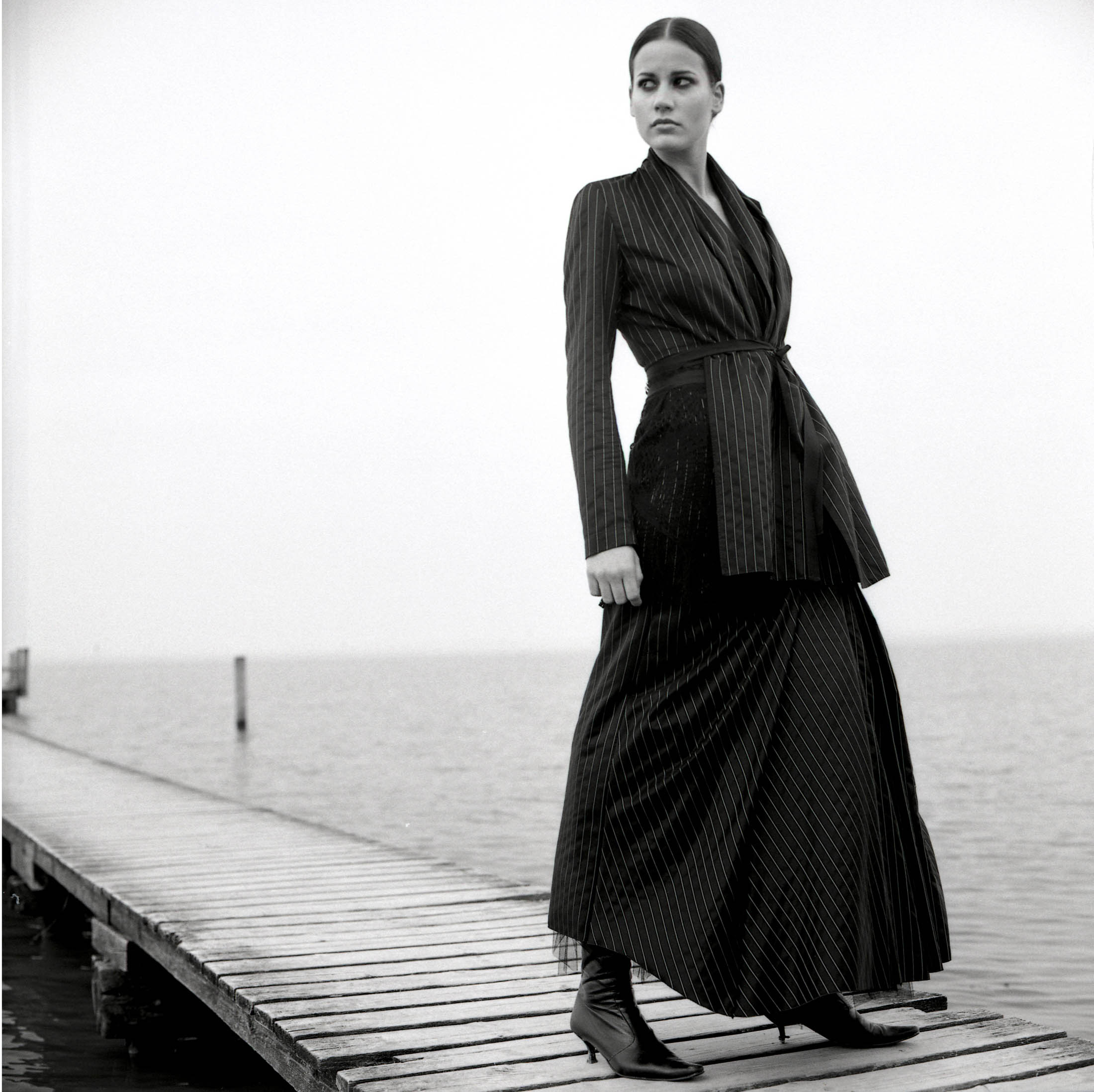 Ein Schwarzweißbild einer Frau, die in einem eleganten Faltenkleid und Stiefeln auf einem Holzsteg steht und mit heiterem Gesichtsausdruck in die Ferne blickt. © Fotografie Tomas Rodriguez