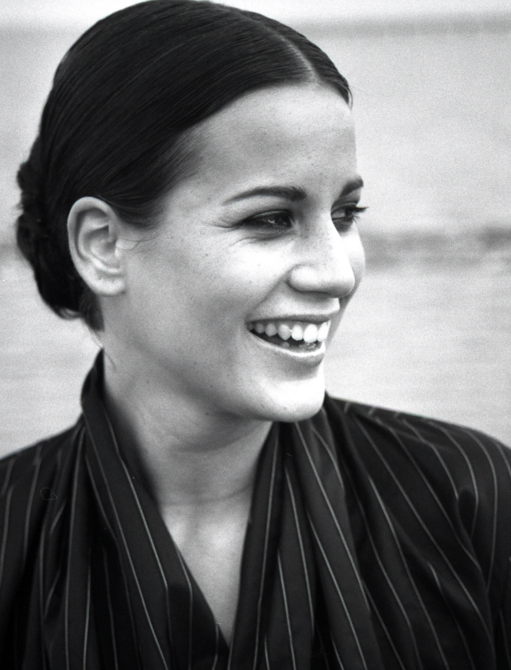Schwarz-Weiß-Porträt einer lächelnden Frau mit zurückgekämmten Haaren, die eine gestreifte Bluse trägt und zur Seite ins Freie blickt. © Fotografie Tomas Rodriguez