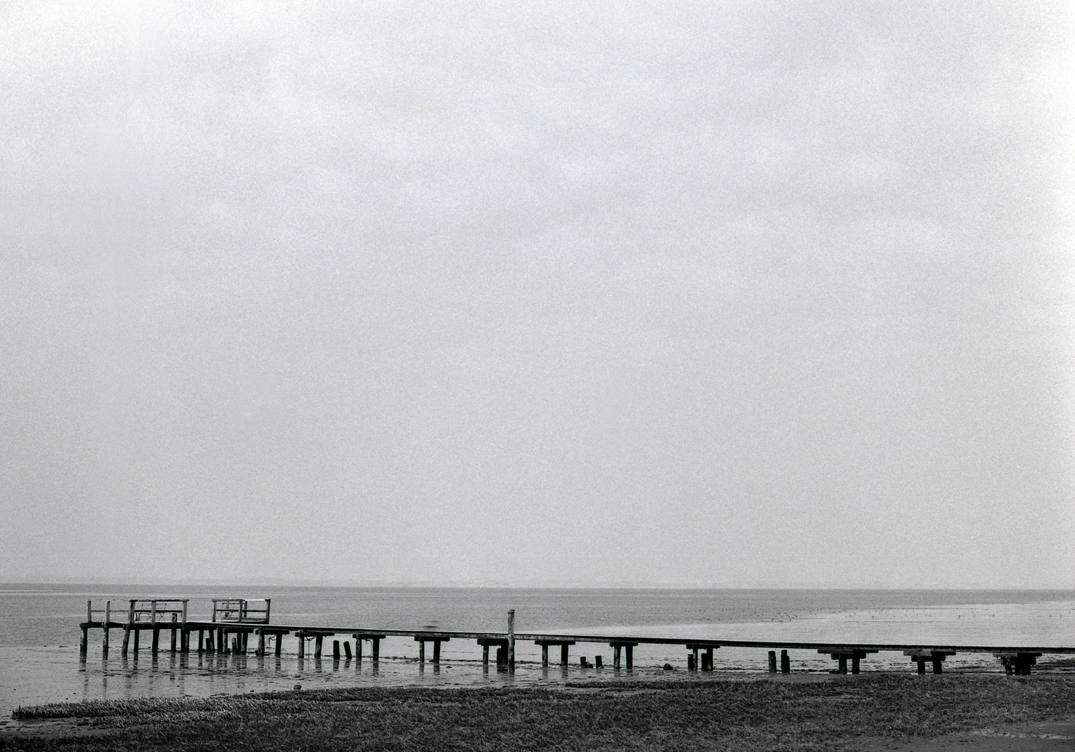 Ein Schwarzweißfoto eines verfallenen Holzstegs, der in einen ruhigen, weitläufigen See unter bewölktem Himmel hineinragt und ein Vintage-Feeling vermittelt. © Fotografie Tomas Rodriguez