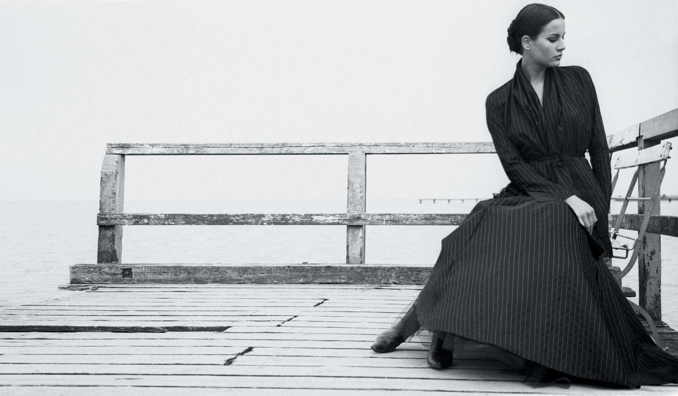 Eine Frau in einem langen, plissierten schwarzen Kleid sitzt nachdenklich auf einem Holzsteg und blickt auf das neblige Meer. Die Stimmung ist nachdenklich und heiter. © Fotografie Tomas Rodriguez