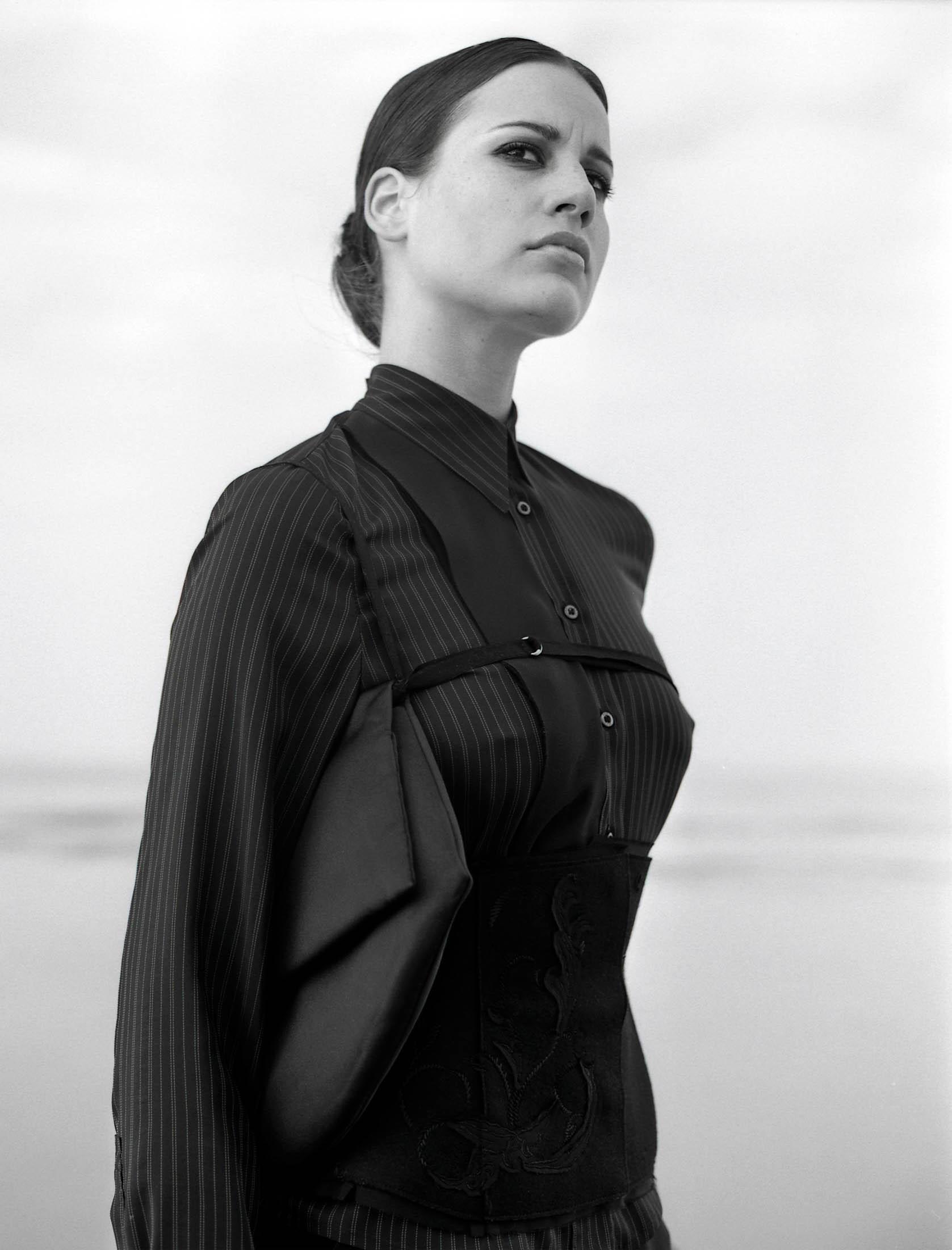 Ein Schwarzweißfoto einer Frau mit zurückgekämmten Haaren, die eine gestreifte Bluse und ein einzigartiges besticktes Korsett trägt und vor einem einfarbigen Hintergrund steht. Sie blickt mit ernster Miene zur Seite. © Fotografie Tomas Rodriguez