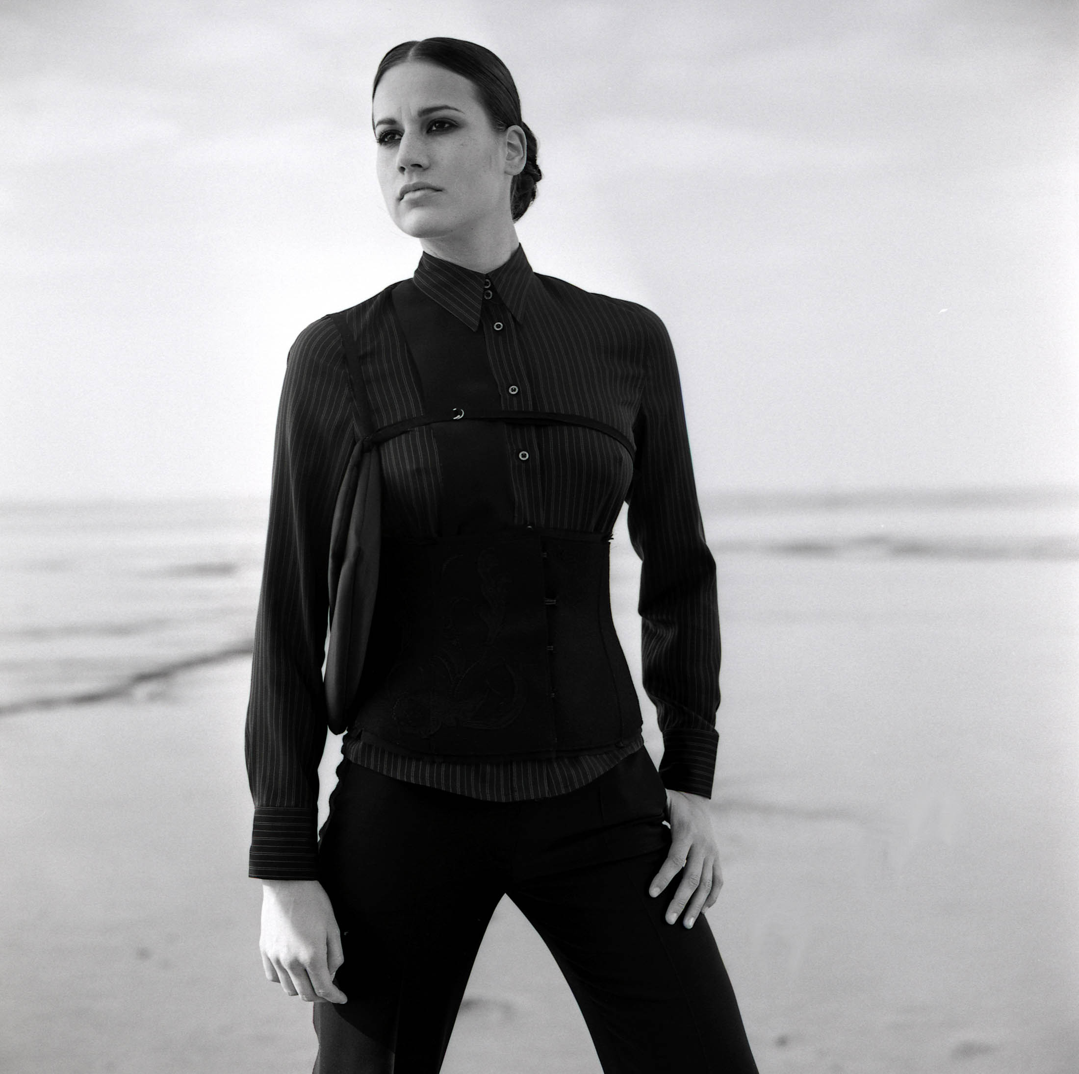 Ein Schwarzweißporträt einer Frau, die an einem Strand steht. Sie trägt eine elegante schwarze Bluse und Hose und blickt mit neutralem Gesichtsausdruck aufmerksam in die Kamera. © Fotografie Tomas Rodriguez